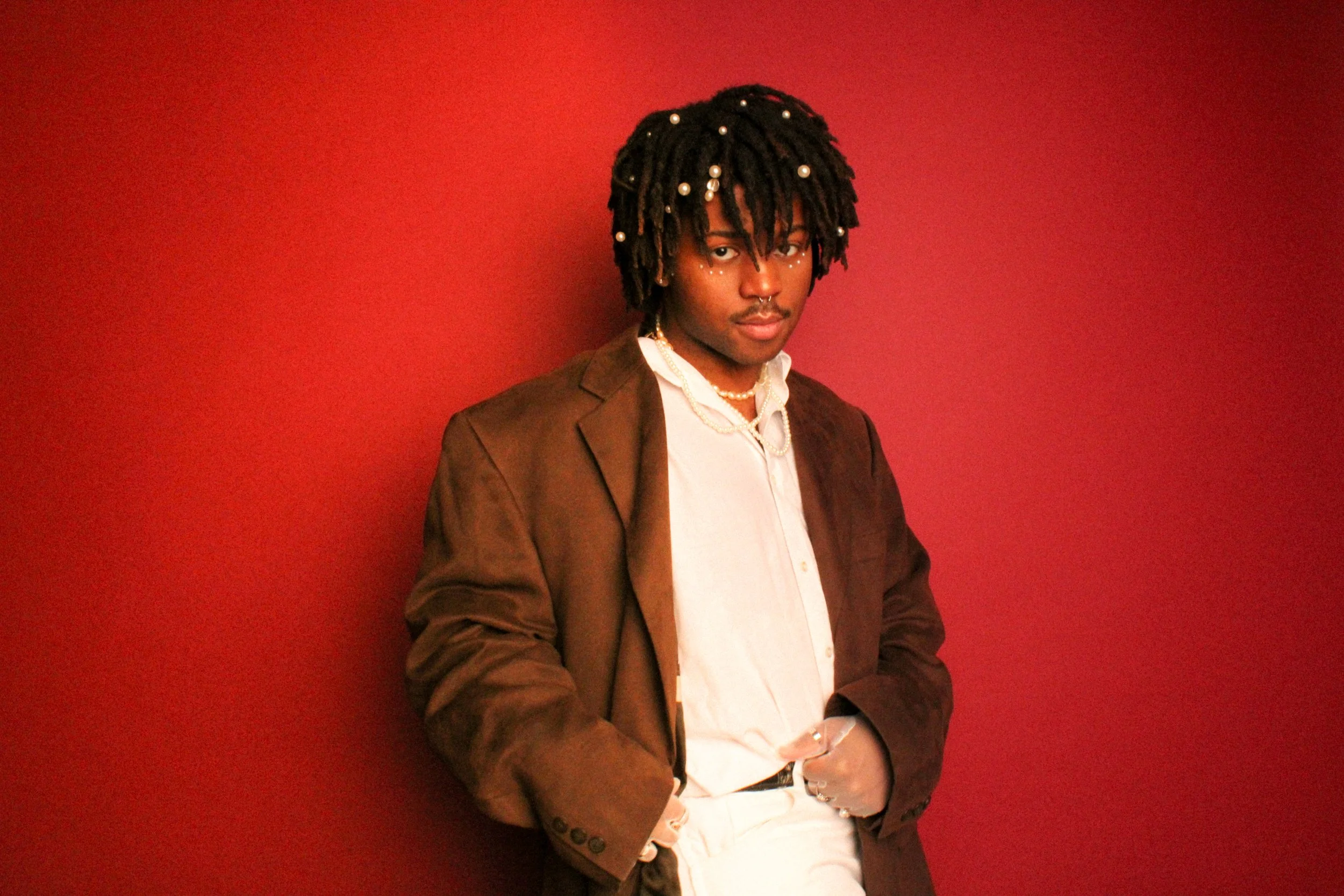 A young man with dreadlocks decorated with beads, wearing a brown blazer, white shirt, and layered necklaces, standing against a red background.