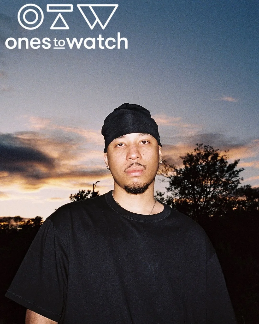 Portrait of a young man wearing a black t-shirt and head covering, standing outdoors during sunset, with trees and a colorful sky in the background, and the 'ones to watch' logo in the top left corner.