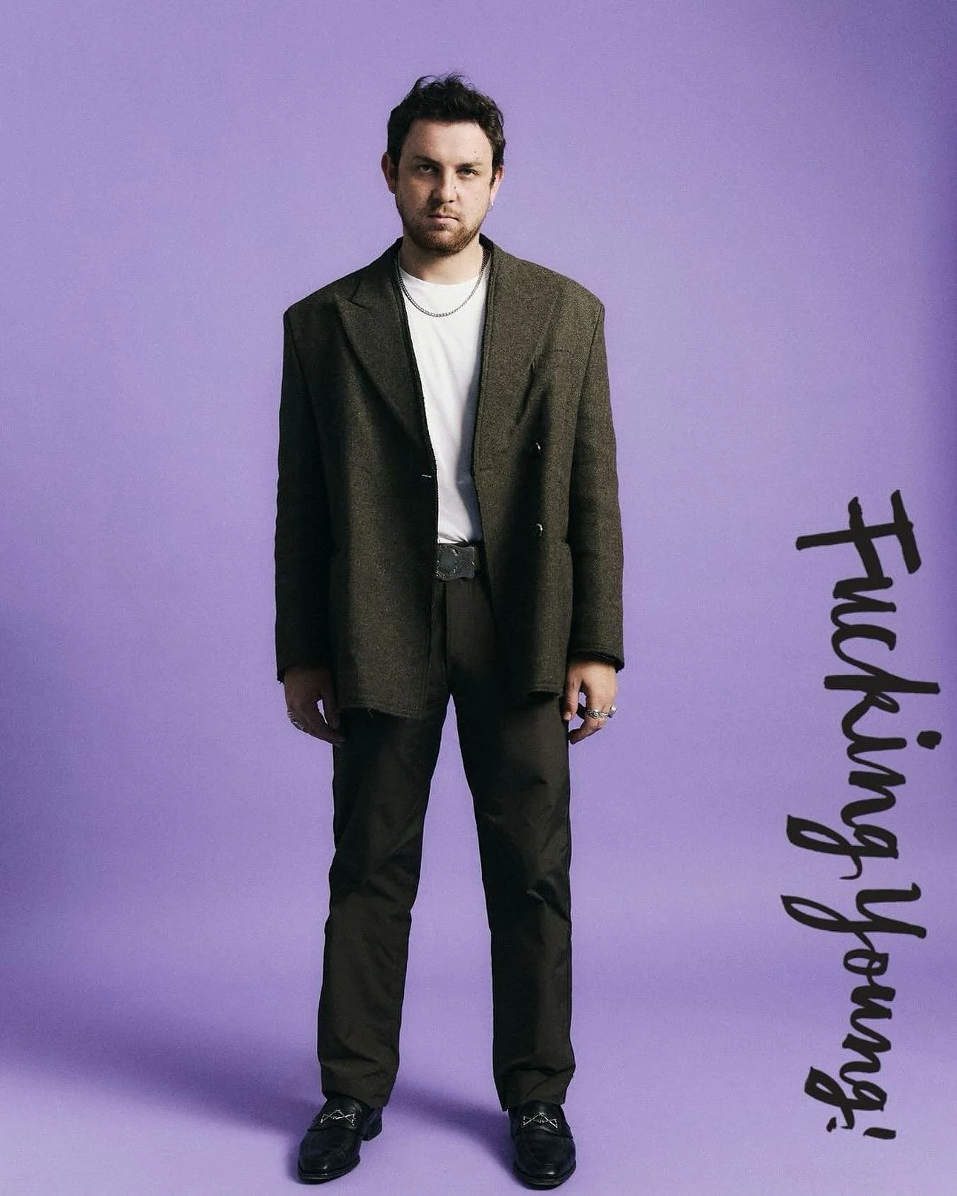 A man standing against a purple background, wearing a dark blazer, white shirt, dark pants, and black shoes. The text 'Fucking ahead!' is written vertically on the right side of the image.