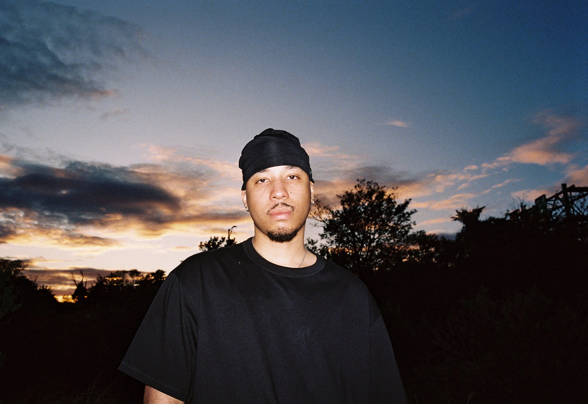 A young man wearing a black beanie and black t-shirt stands outdoors during sunset, with trees and a partly cloudy sky in the background.