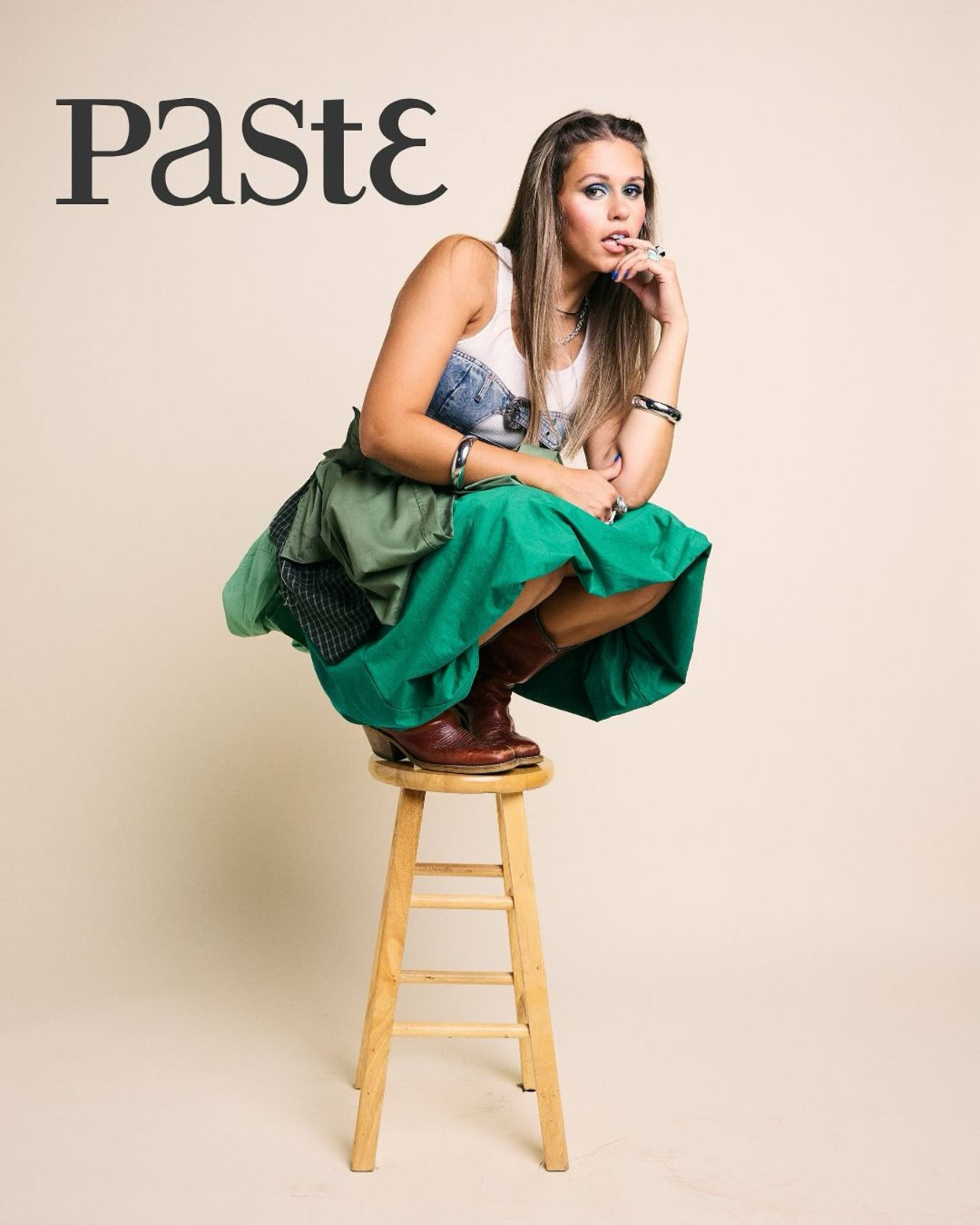 Young woman crouching on a stool against beige background, wearing a sleeveless top, long skirt, and brown boots, with the word 'PASTE' in stylized black font at the top left.