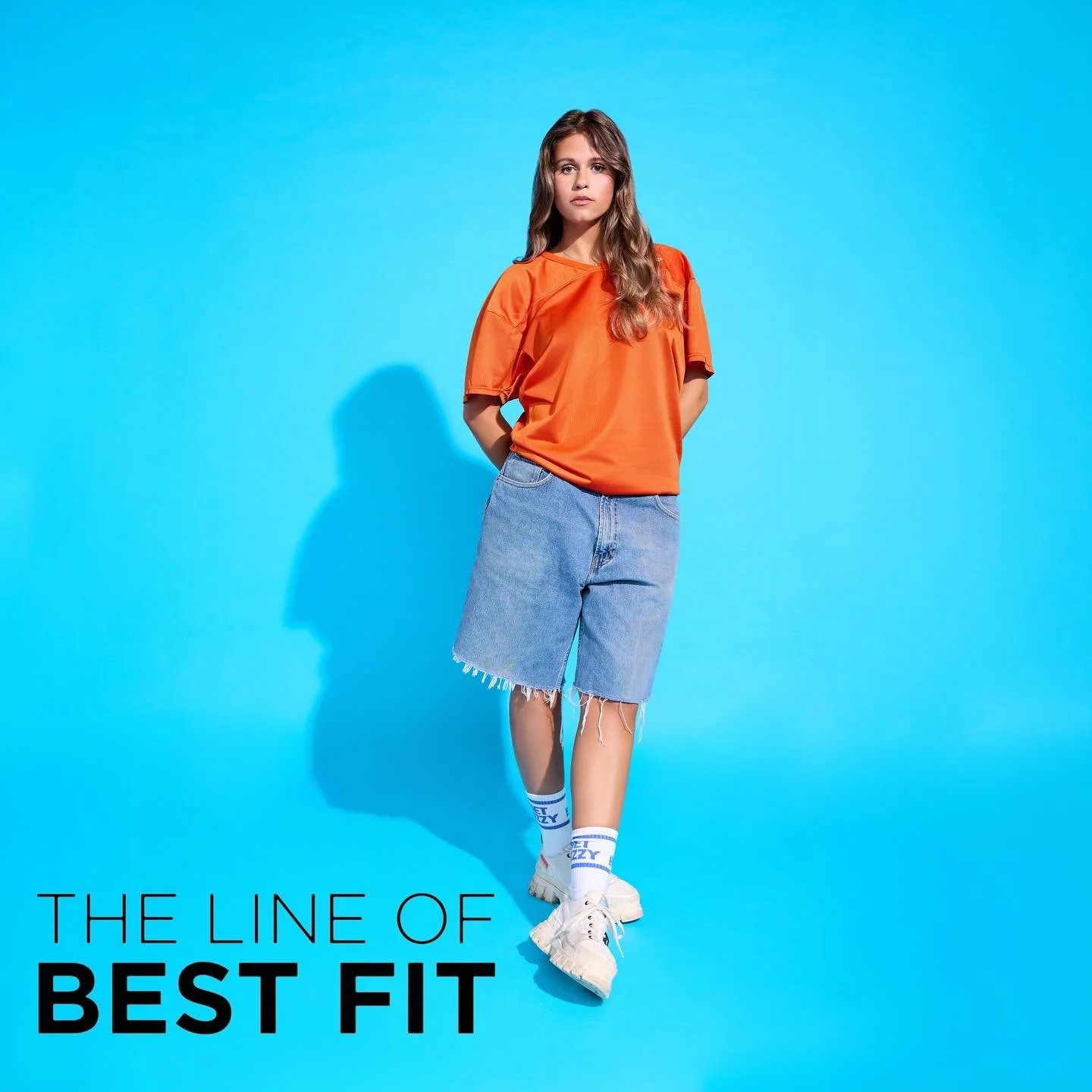 A young woman with long brown hair wearing an orange T-shirt, denim shorts, white sneakers, and white socks standing against a blue background, with the text 'The Line of Best Fit' in the lower left corner.