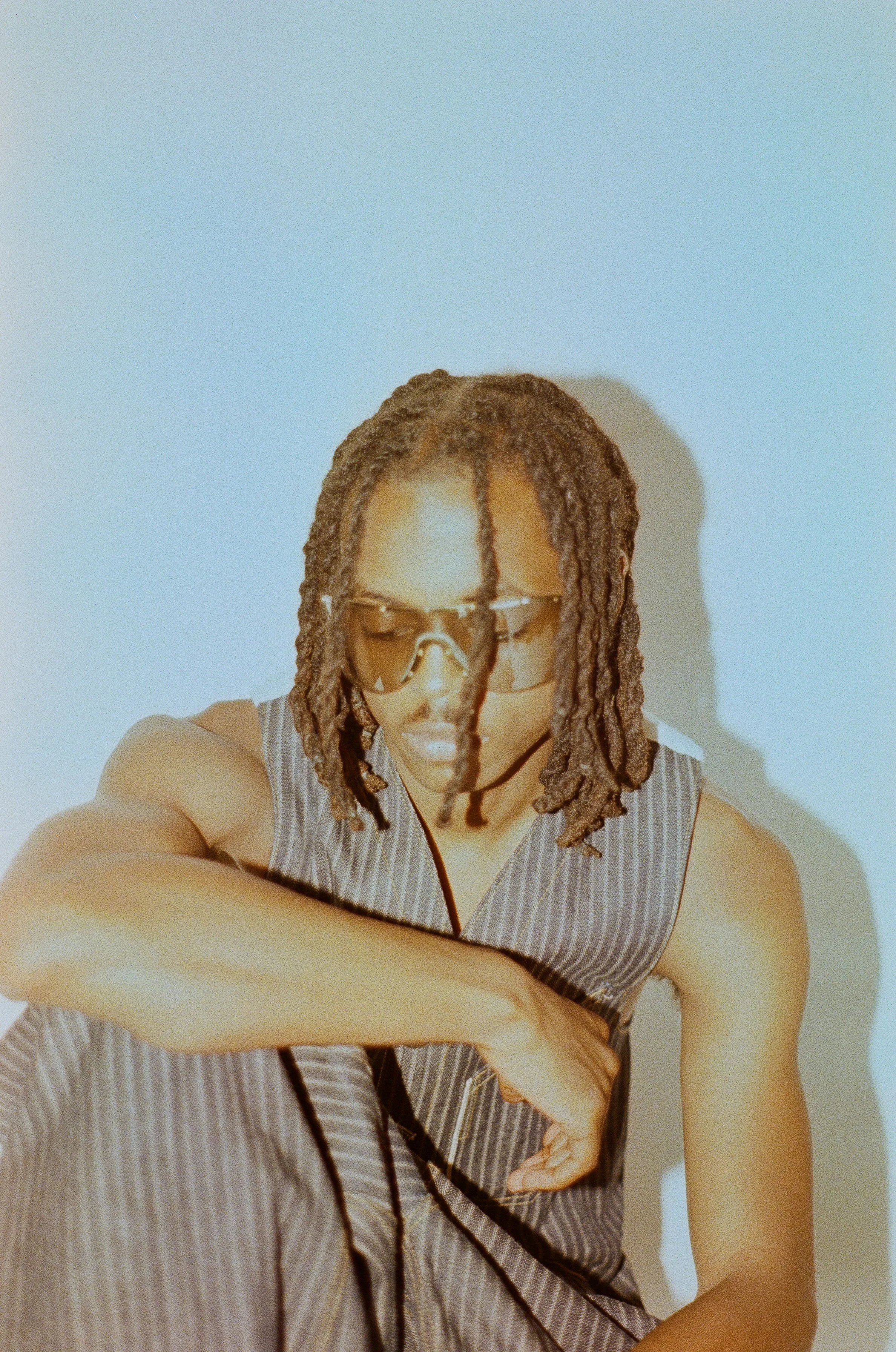 A person with shoulder-length dreadlocks wearing sunglasses and a sleeveless striped shirt, looking down with one arm across their chest, against a plain light-colored background.