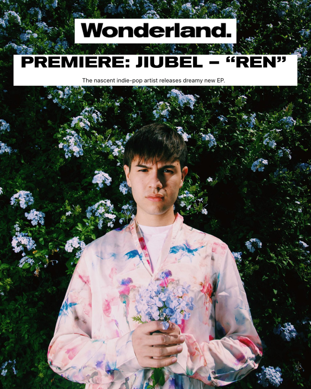 Young man standing in front of a bush with blue flowers, holding a small bouquet of blue flowers, wearing a pastel-colored floral shirt, with a serious facial expression.