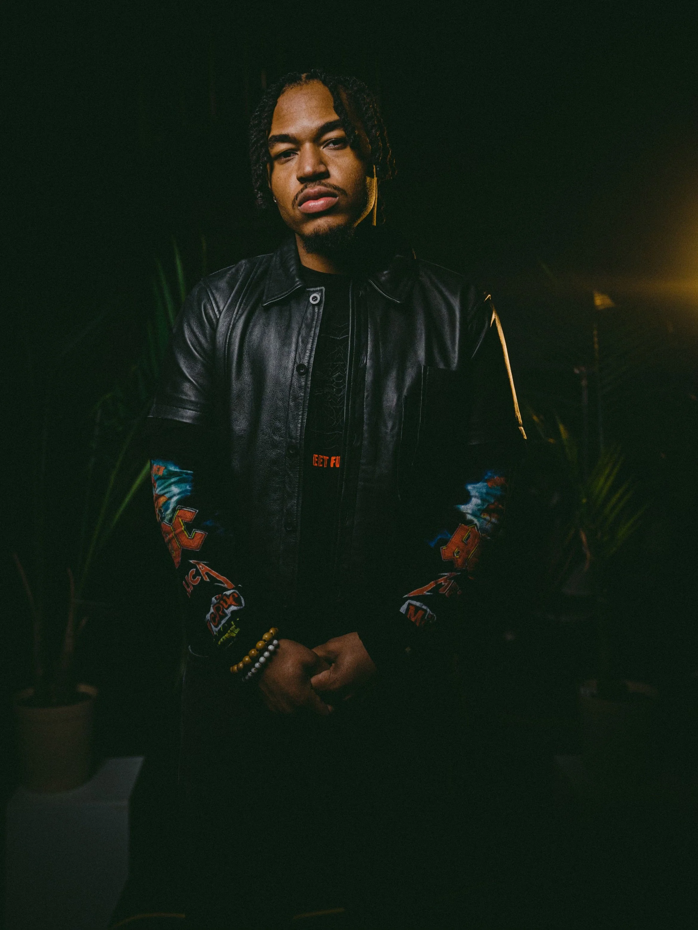 A young man with dreadlocks wearing a black leather jacket and colorful sleeves standing in a dark environment with some plants and lighting behind him.