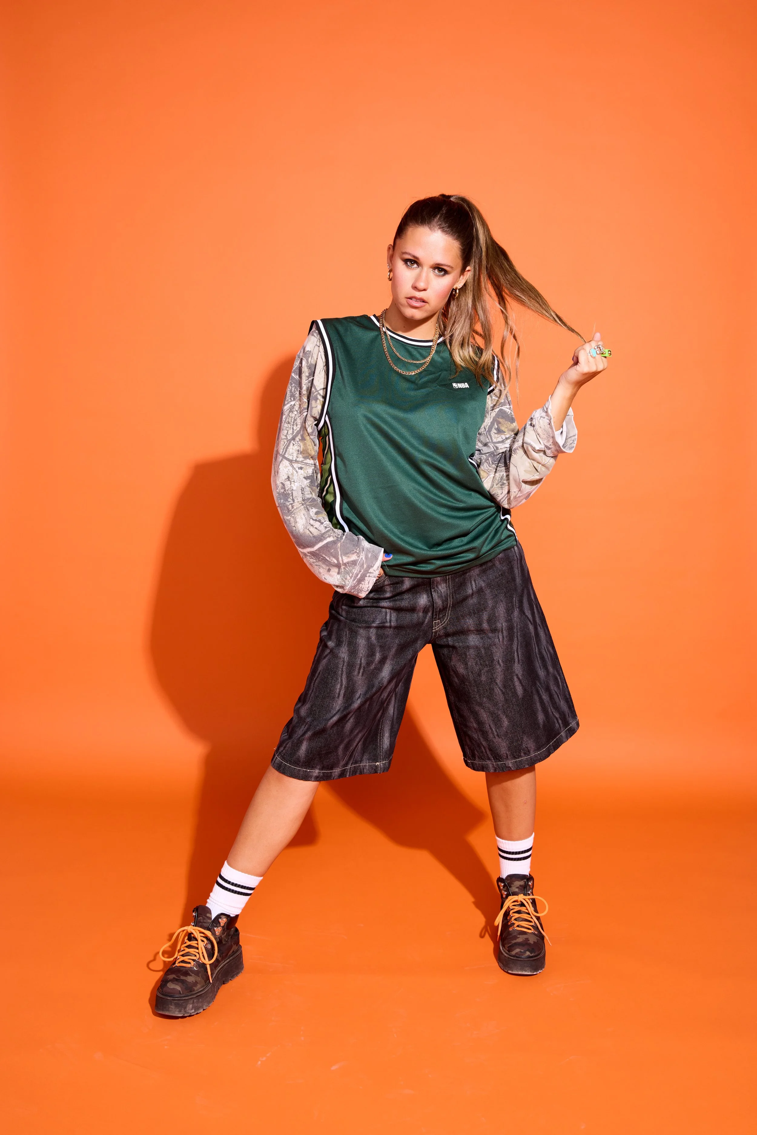 A young woman with a high ponytail wearing a green sports vest over a long sleeve shirt with a gray pattern, black shorts, white socks with black stripes, and black sneakers with orange laces. She is posing against an orange background, with one hand pulling her hair and the other in her pocket.