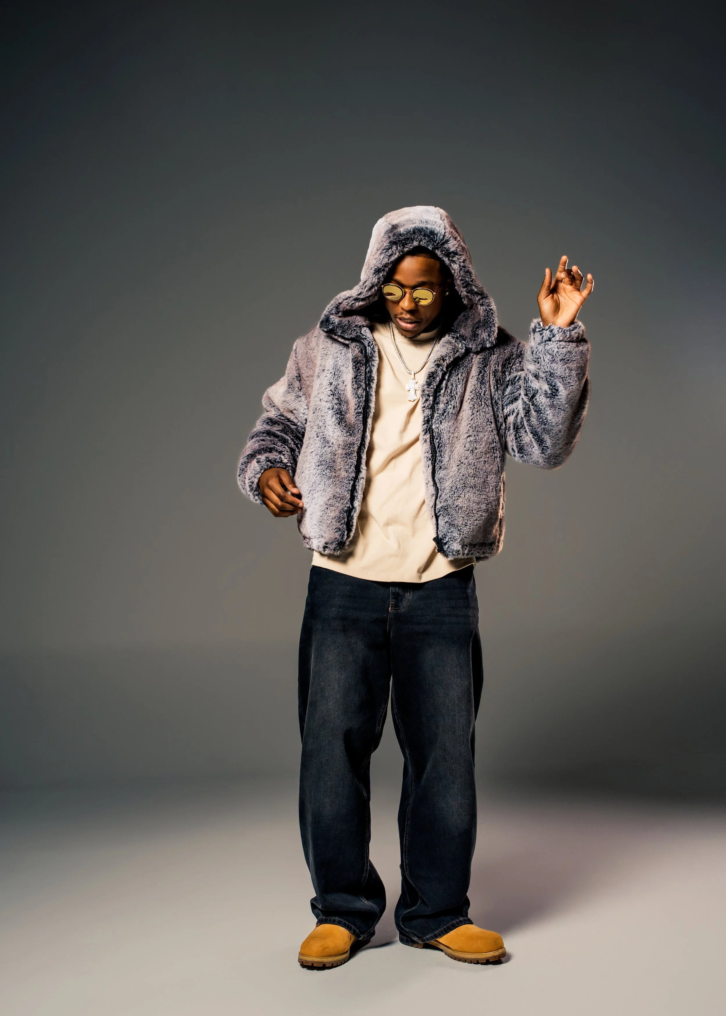 Person wearing a furry jacket, sunglasses, and tan boots, standing against a plain background.