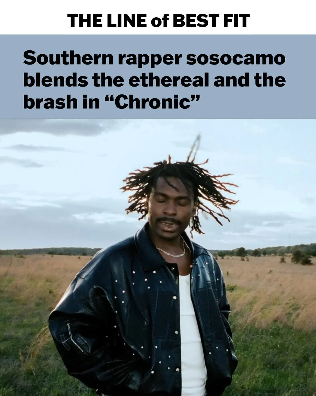 A man with dreadlocks wearing a black leather jacket stands outdoors in a field, with a mostly cloudy sky in the background.