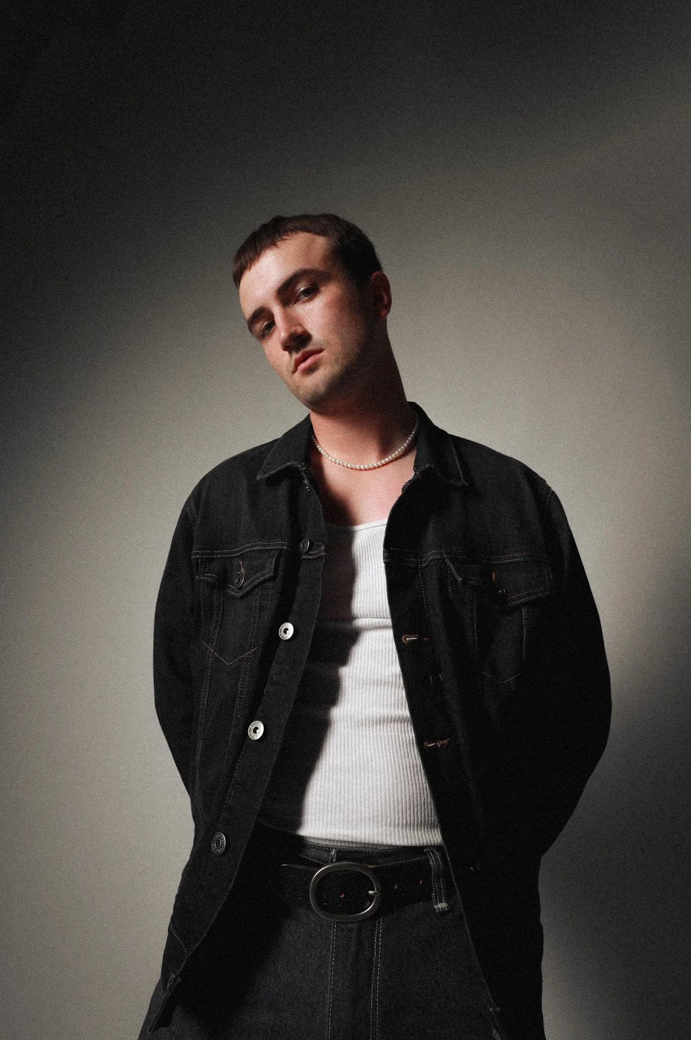 Young man with dark hair in a black denim jacket, white tank top, and black jeans, wearing a pearl necklace, standing against a gradient gray background.