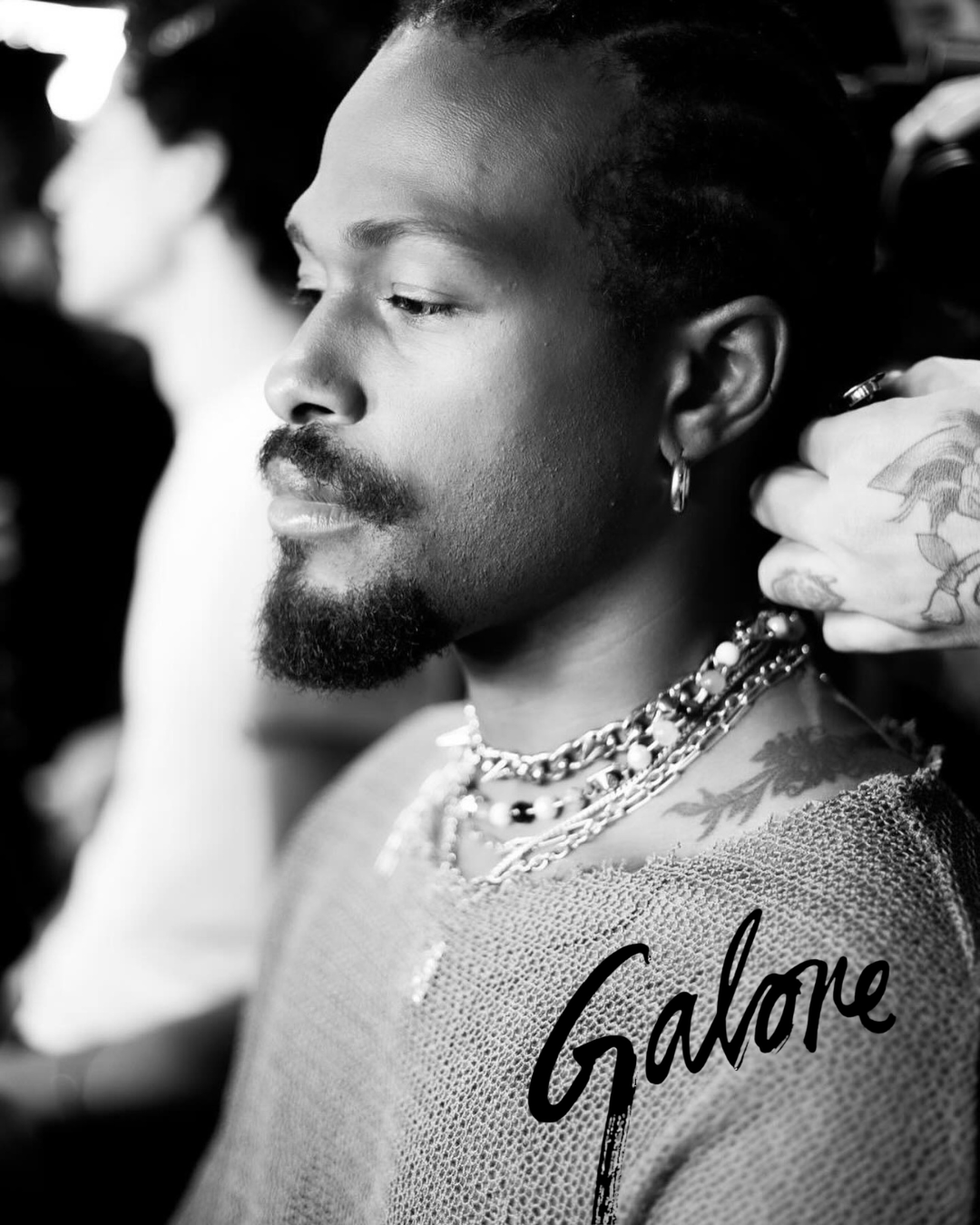 Close-up black and white photo of a man with facial hair and tattoos, wearing layered necklaces and a textured shirt, getting styled or dressed with a hand adjusting his earring.