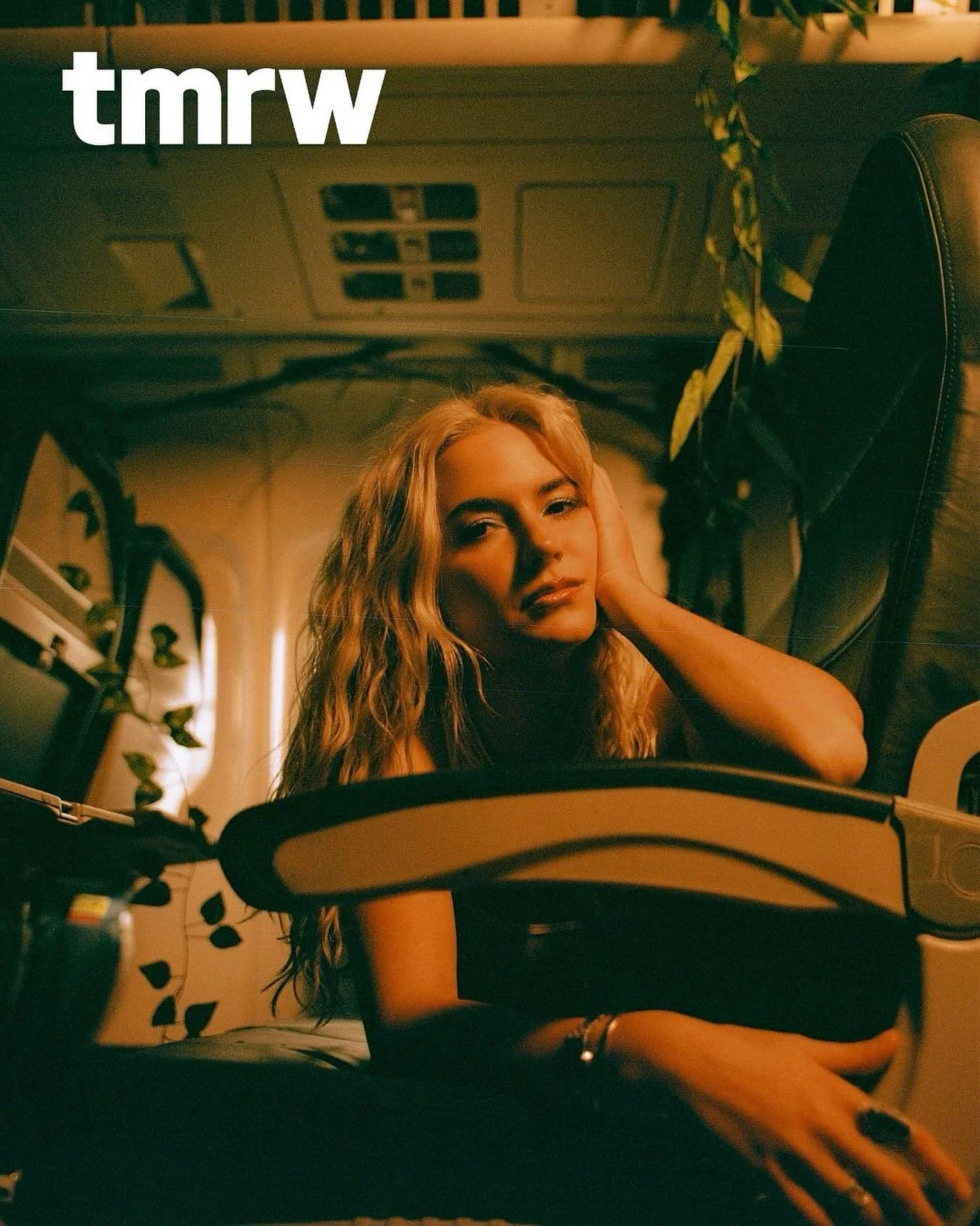 A woman with blonde, wavy hair lying inside a car, resting her head on one hand, with a soft, warm lighting in the scene.