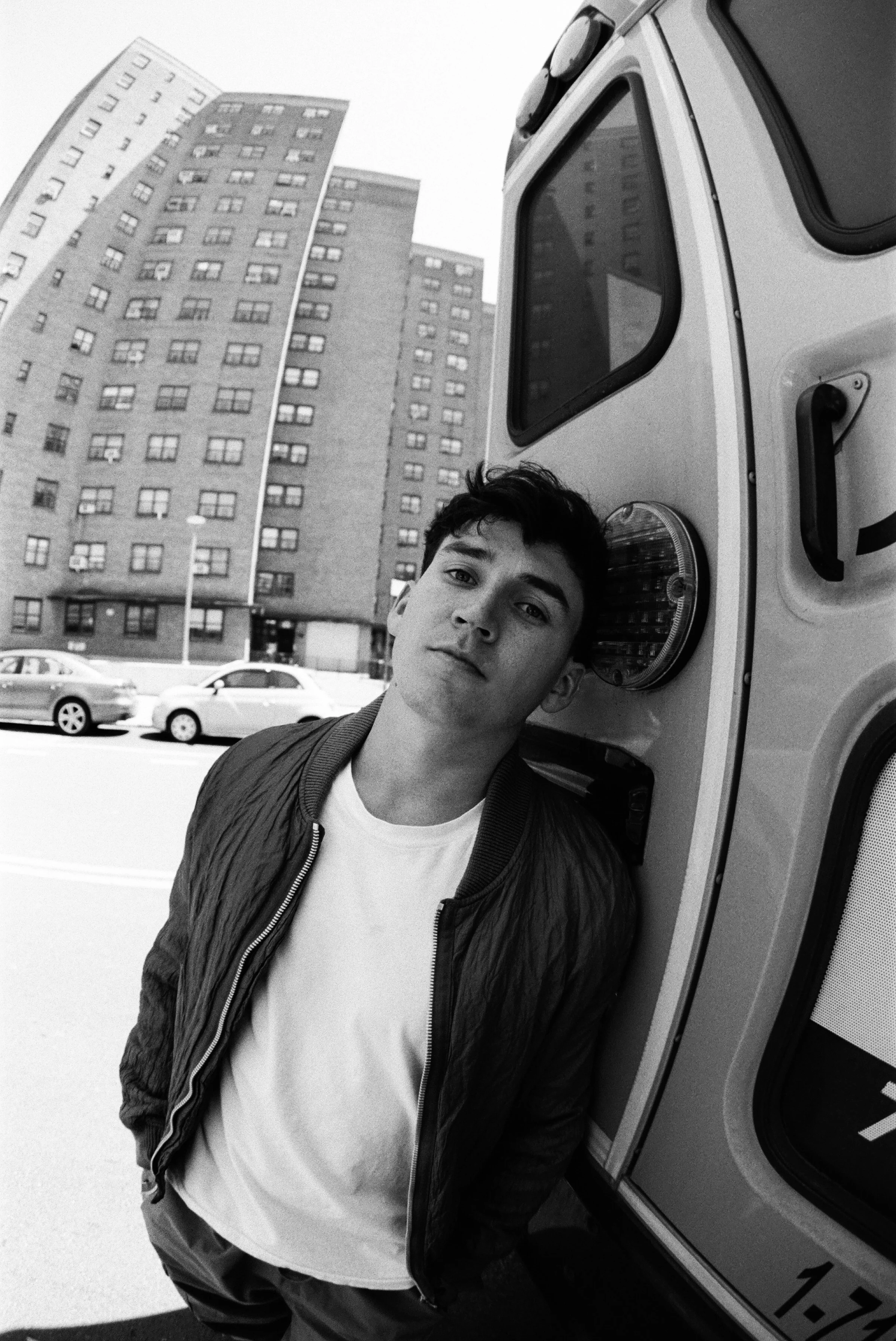 A young man with dark hair leaning against an ice cream truck on a city street, with tall apartment buildings and parked cars in the background, in black and white.