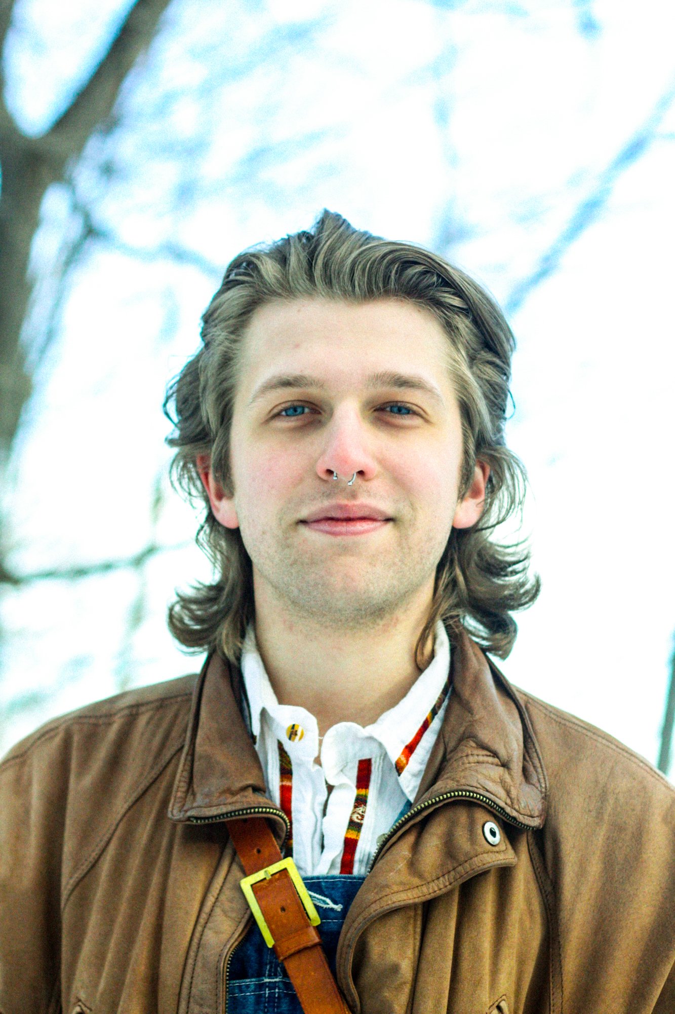 A young man with long, wavy brown hair, wearing a brown jacket, white shirt, and overalls, standing outdoors with trees and a cloudy sky in the background.
