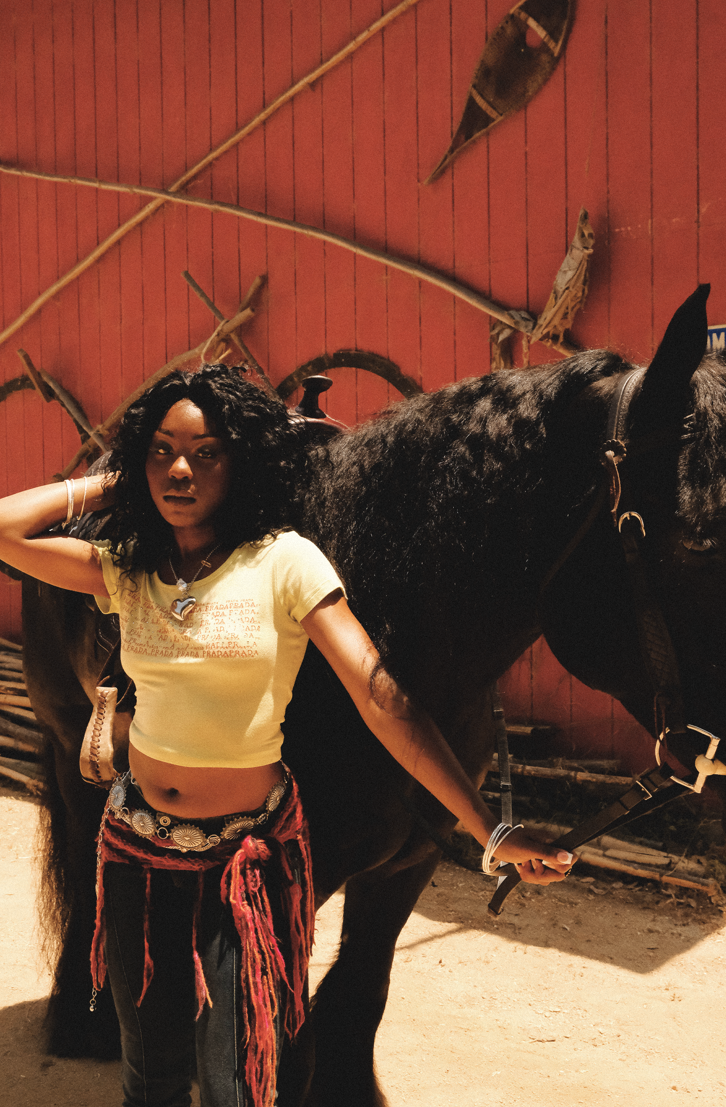 A young woman with curly hair, wearing a yellow crop top and a colorful skirt, stands next to a black horse against a red wall with decorative boomerangs and sticks.