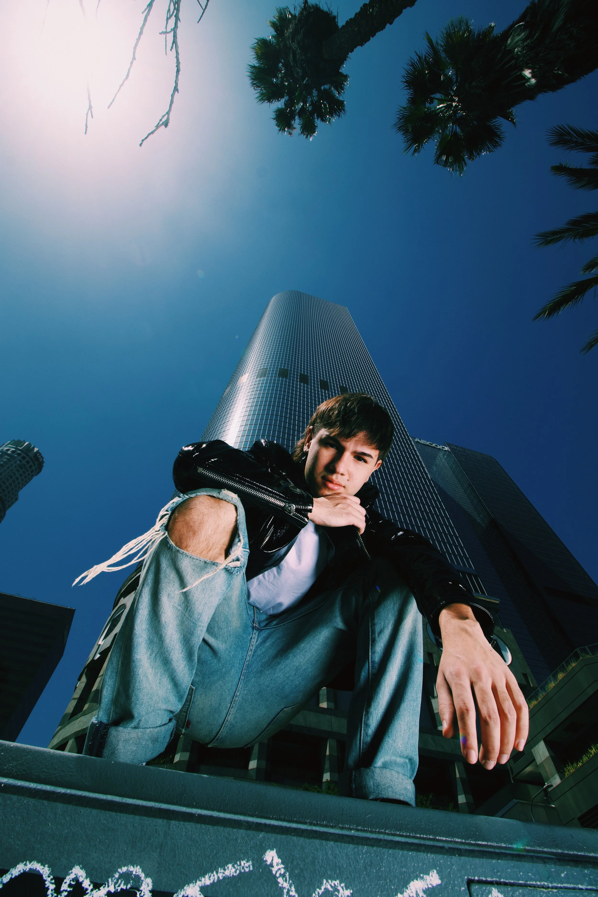 Young man crouching in front of a tall, modern skyscraper with palm trees nearby, under clear blue sky.