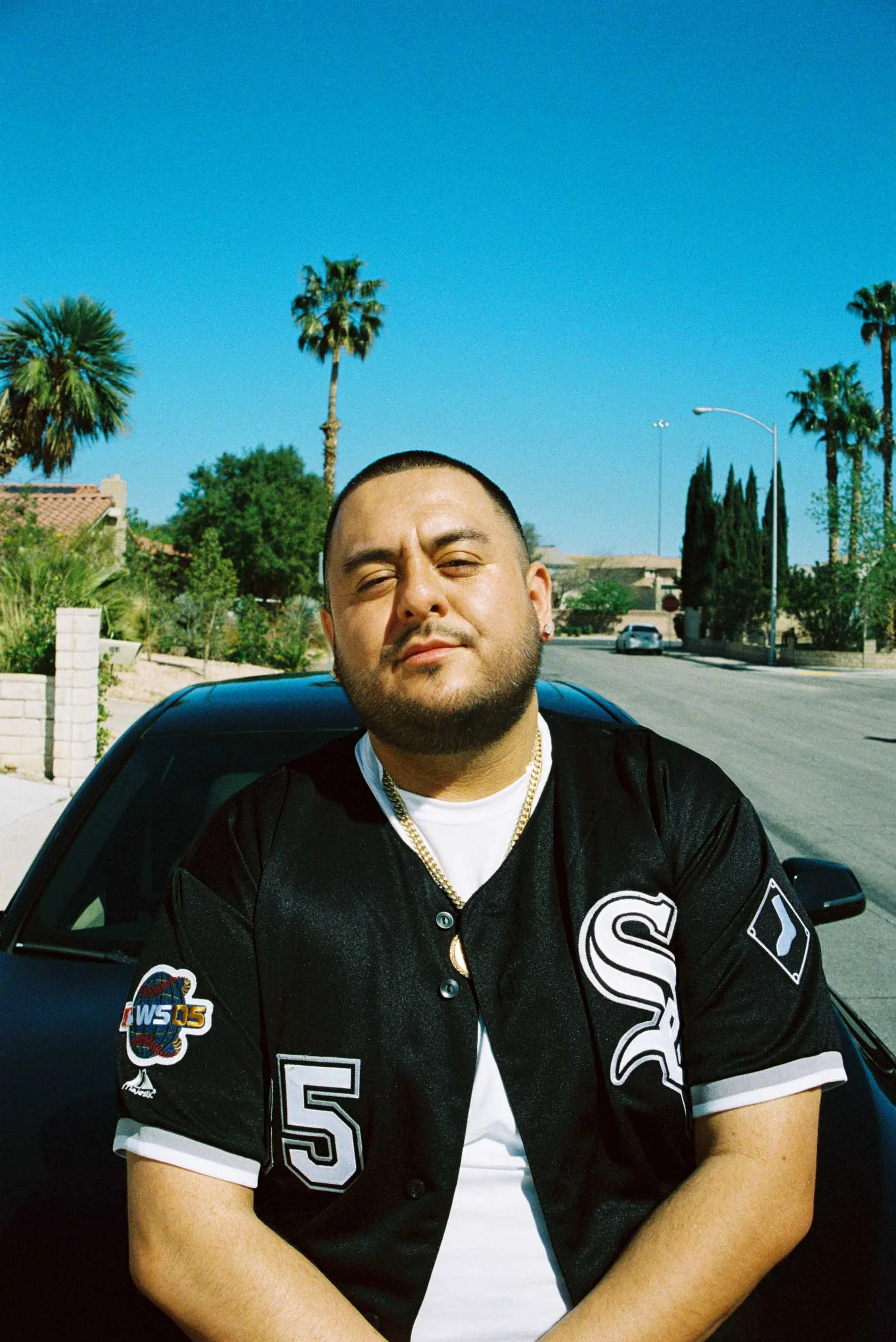 A man with a beard and short hair sitting in front of a black car on a sunny day, wearing a black Chicago White Sox baseball jersey and gold chains.