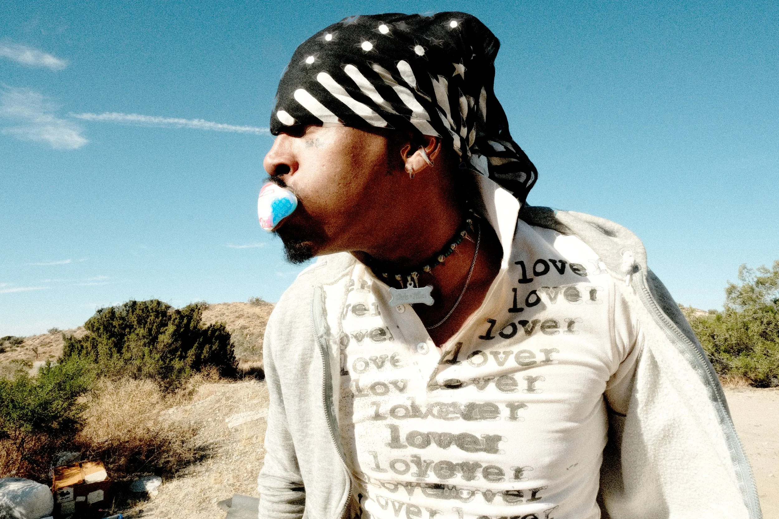 A man with dark skin wearing a black and white bandana over his eyes, blowing a bubble with bubblegum, outdoors in a desert landscape with bushes and a clear blue sky.