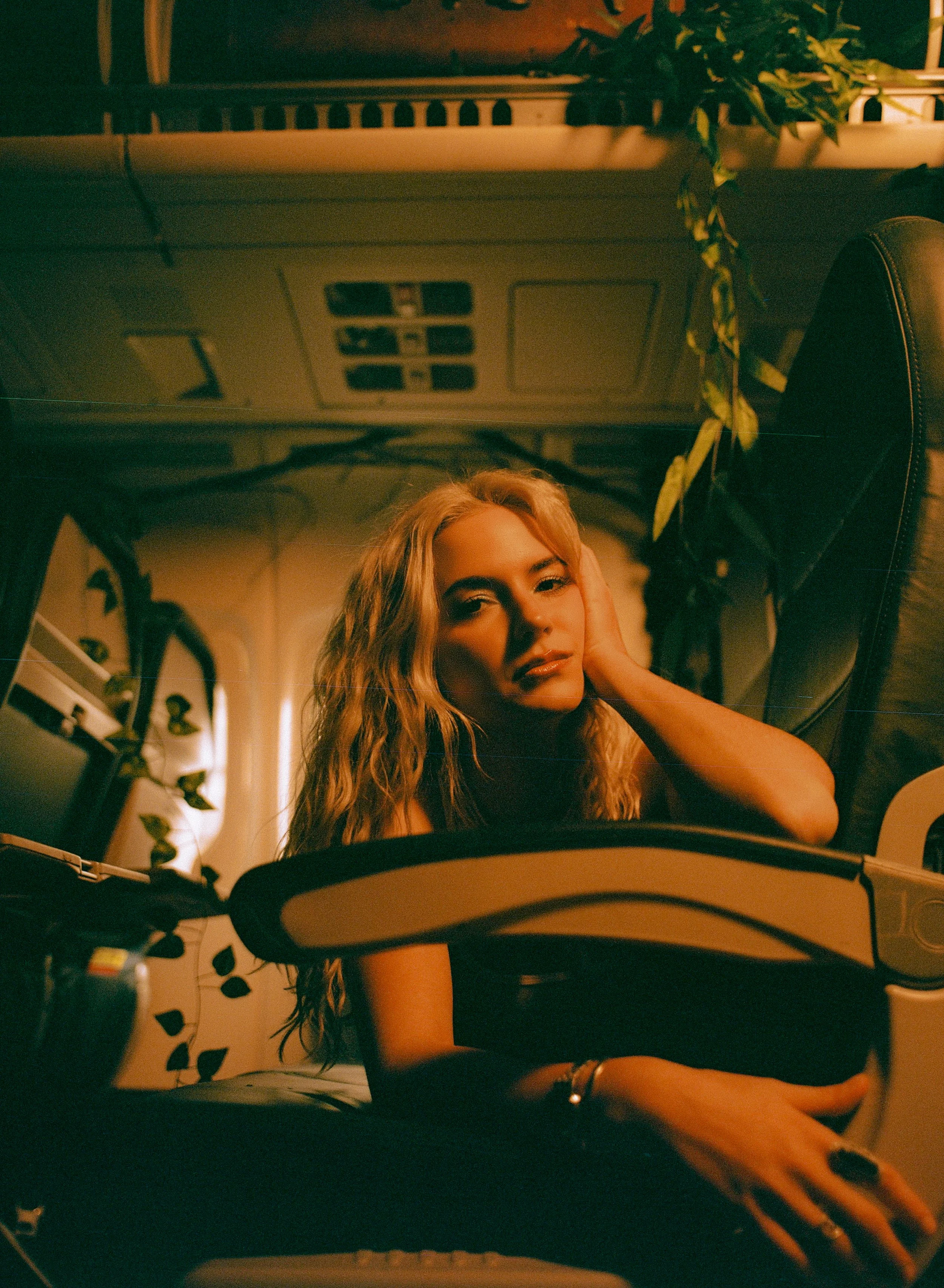 A young woman with curly blonde hair sitting inside a vehicle, looking at the camera with a relaxed expression, illuminated by warm, orange lighting.