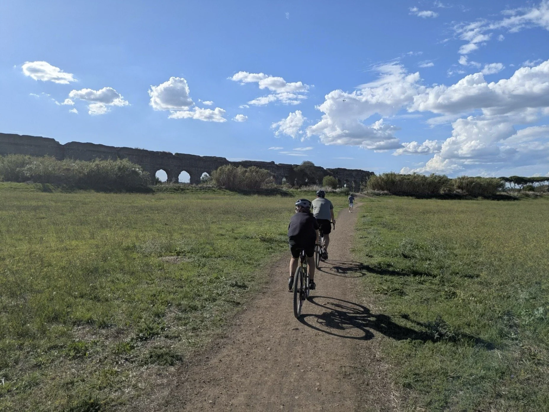 Cyclists riding on a dirt path through a grassy field, with an ancient Roman aqueduct in the background under a partly cloudy sky.