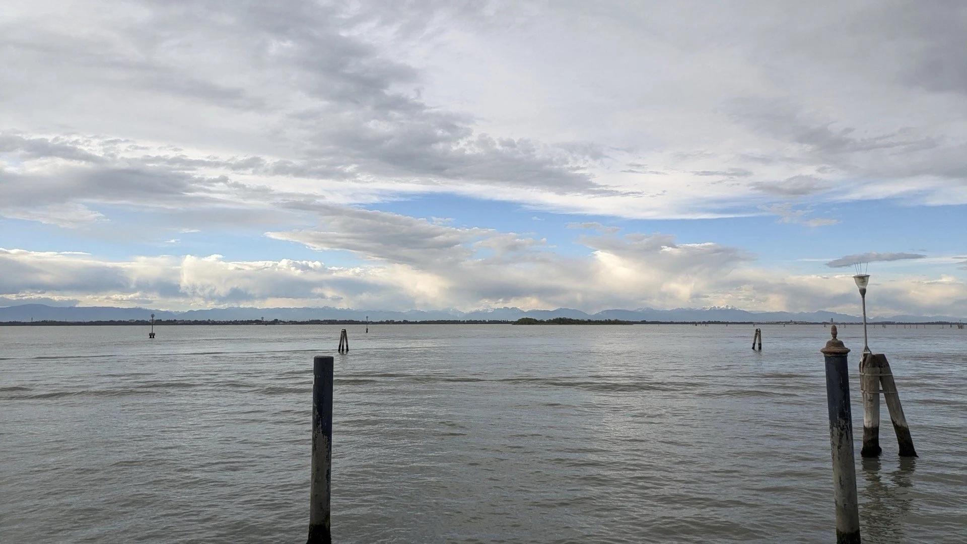 Overcast sky above a calm body of water with distant mountains and small islands on the horizon. Several weathered wooden posts and a lamp post are visible in the water.