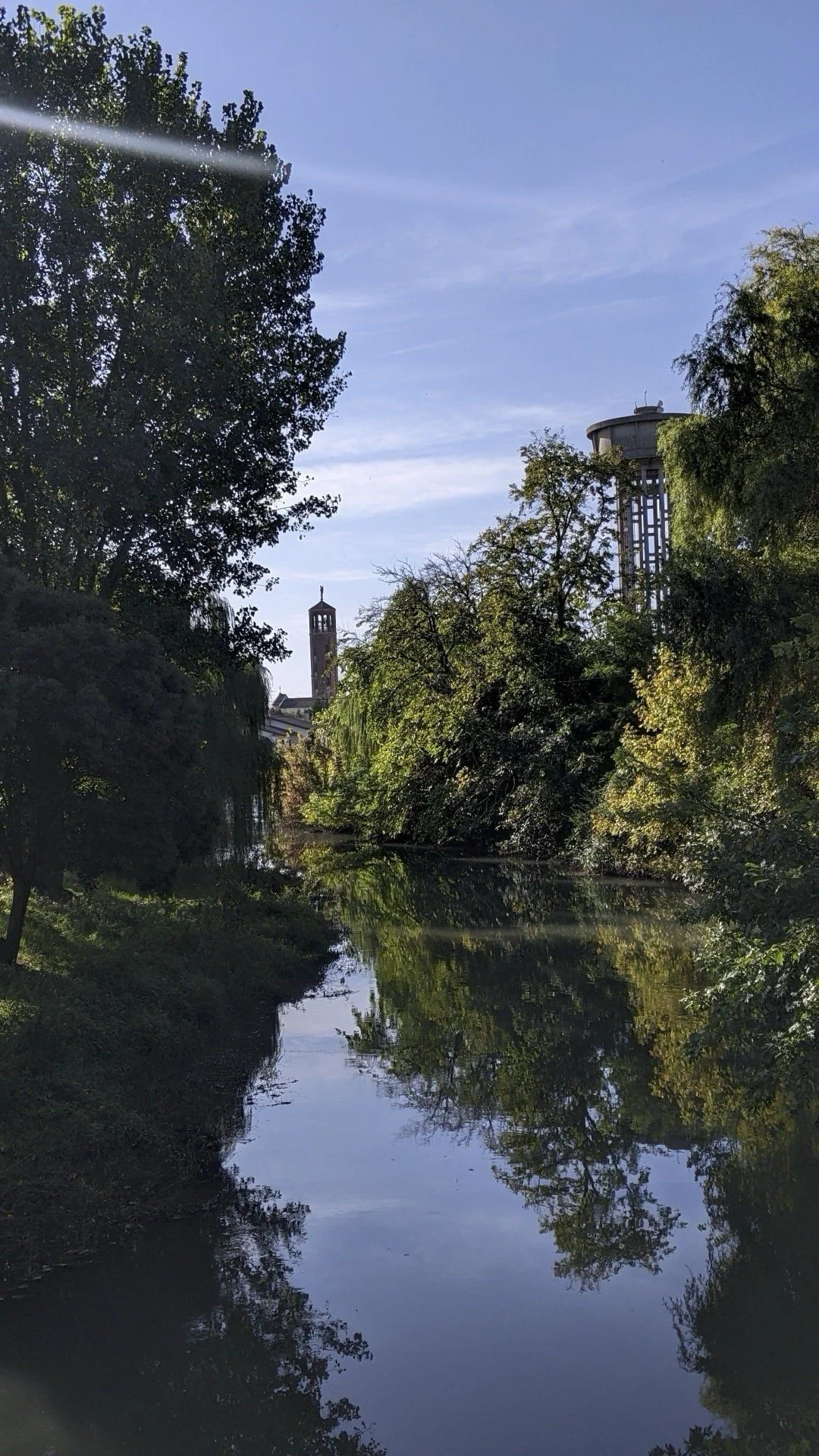 A calm river reflecting the trees along its banks, with a tall building and a tower visible in the background under a blue sky.