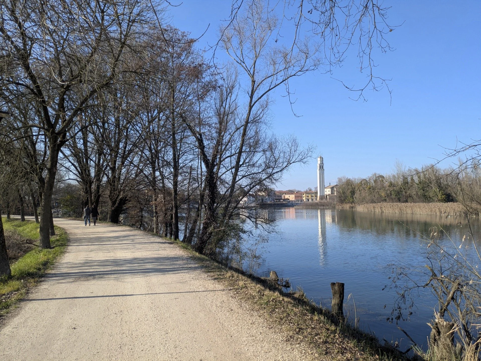 A dirt walking path runs alongside a river with a church or bell tower in the distance under a bright blue sky, with trees lining the path and some in the water.