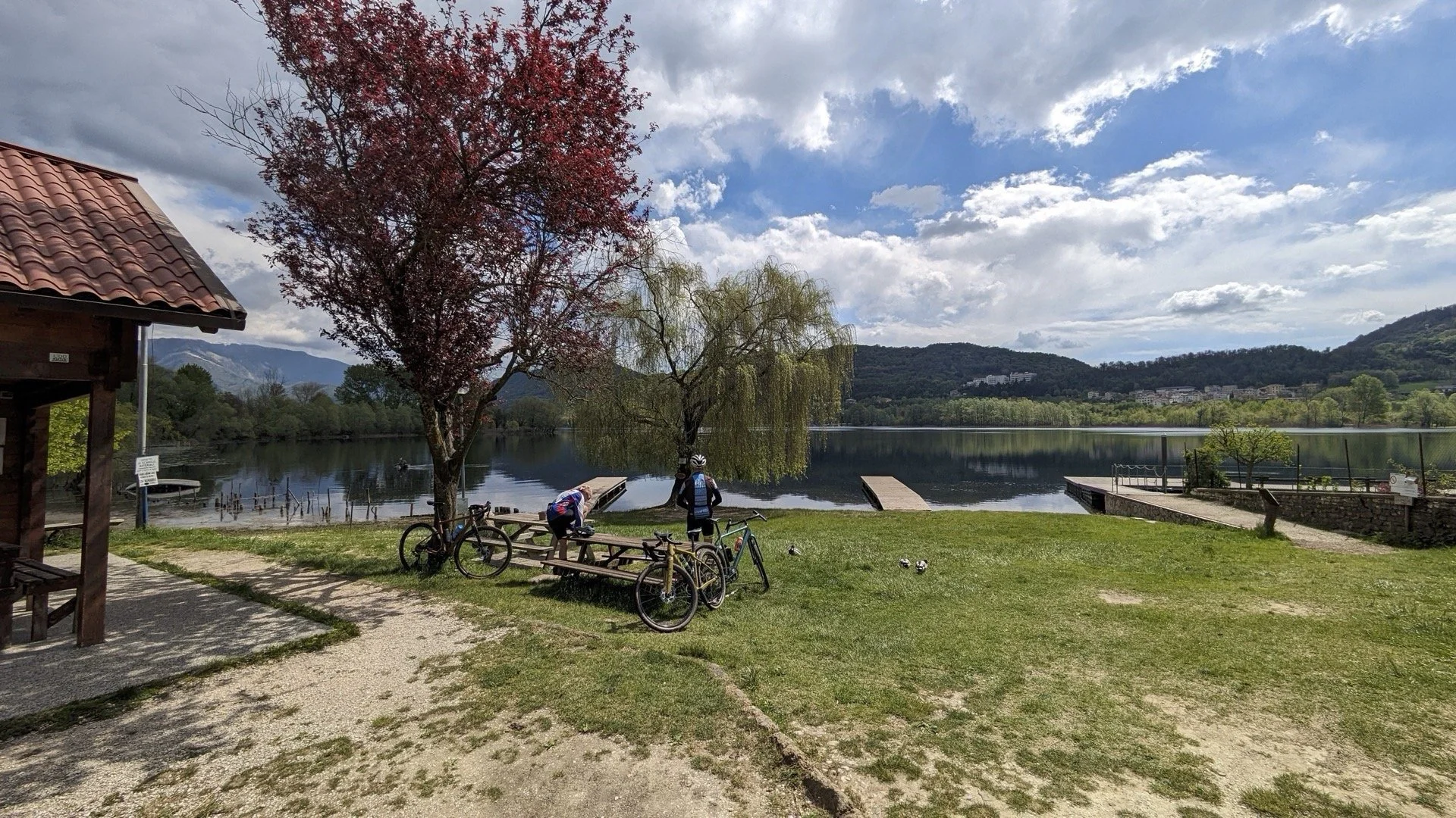 View of a lakeside park with two children and a woman, two bicycles, benches, trees, and a dock under a partly cloudy sky, with mountains in the distance.