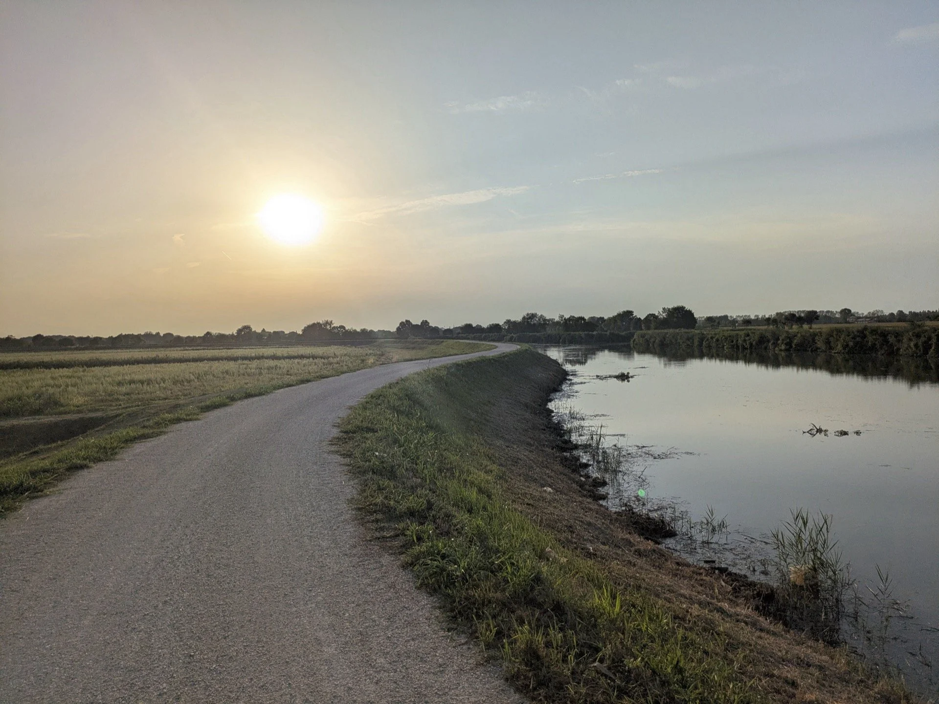 A gravel path runs alongside a calm river with grassy banks, under a setting sun in a rural landscape.