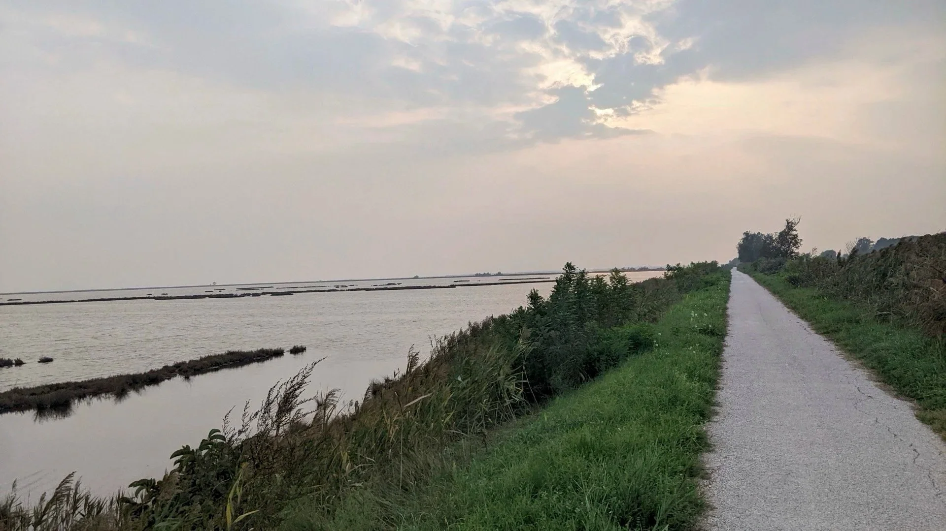 A narrow gravel path runs alongside a body of water, with green grass and shrubs on either side, under a partly cloudy sky.