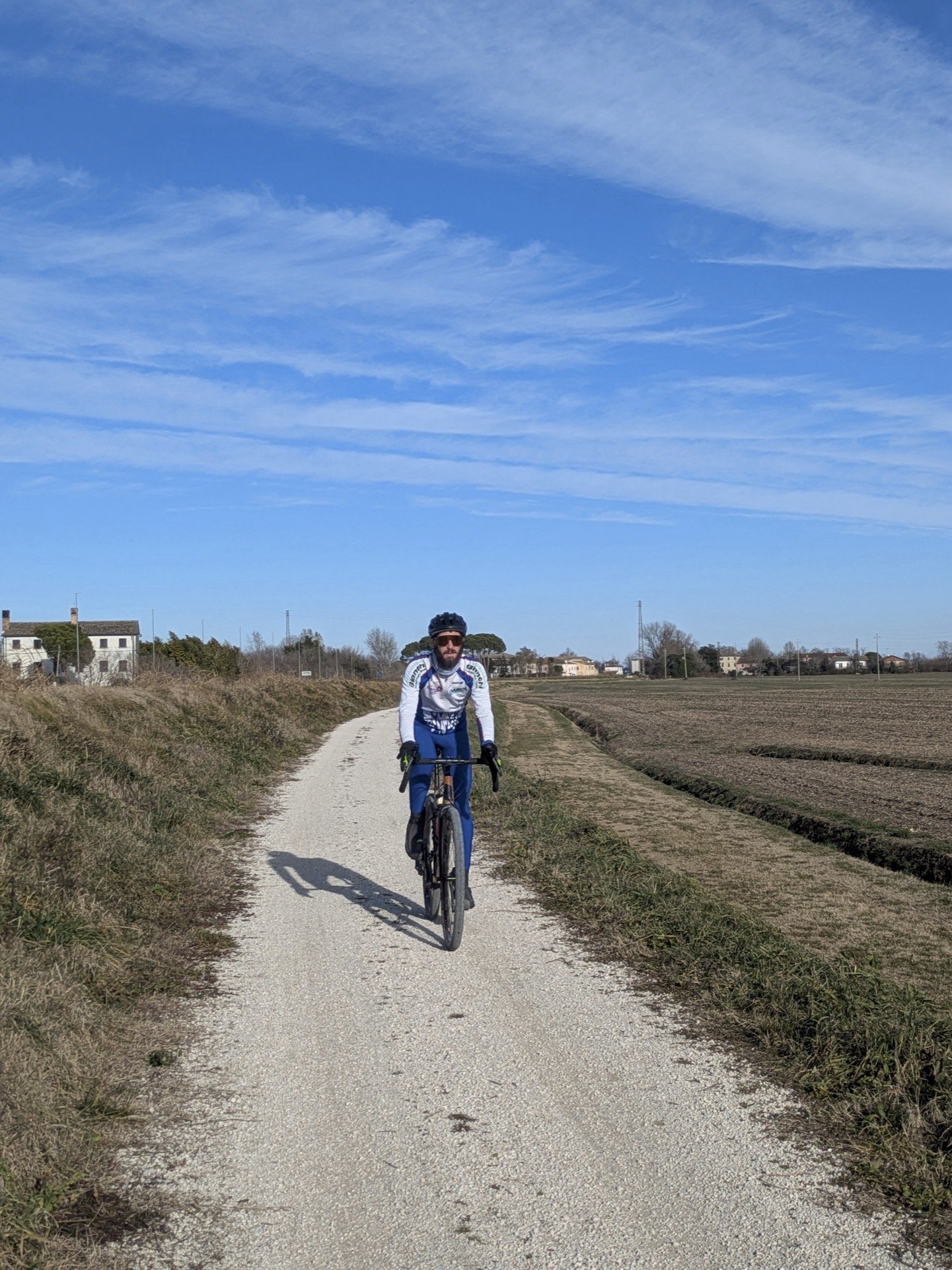 A man riding a mountain bike on a dirt trail through a rural landscape with houses in the distance and a blue sky with wispy clouds.