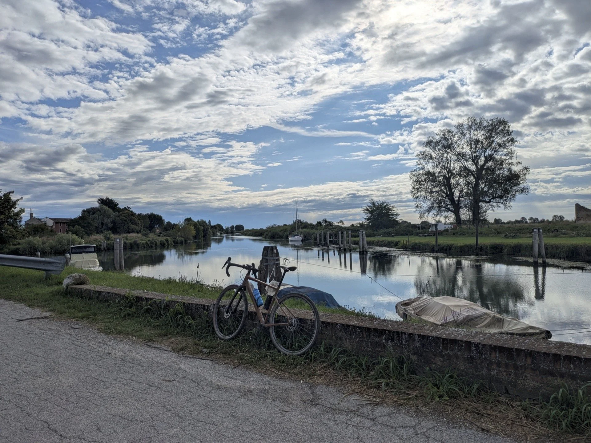 A canal with boats docked alongside, a bicycle leaning against a concrete block, and a tree with sparse leaves under a partly cloudy sky.