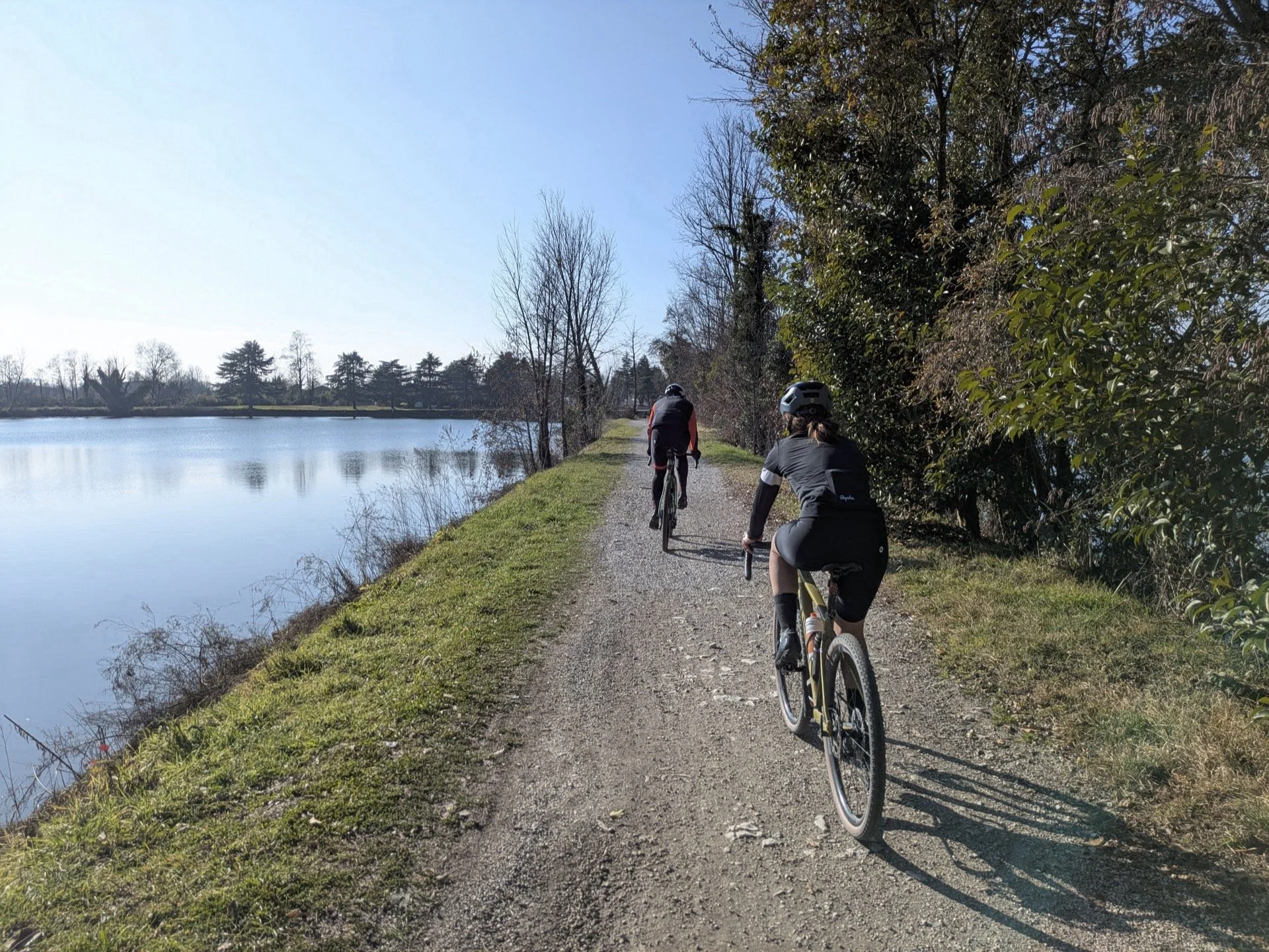 Three people riding bicycles on a gravel path beside a lake, with trees on the right side and clear blue sky.