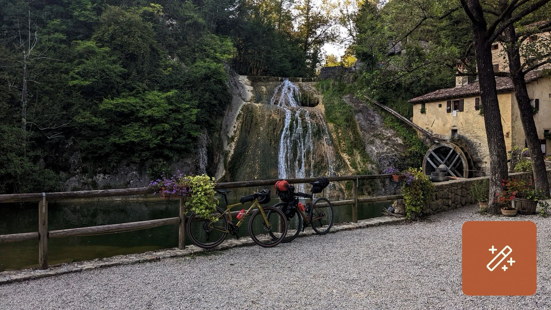 Two bicycles parked against a wooden fence beside a small river and waterfall, with an old building and trees in the background.