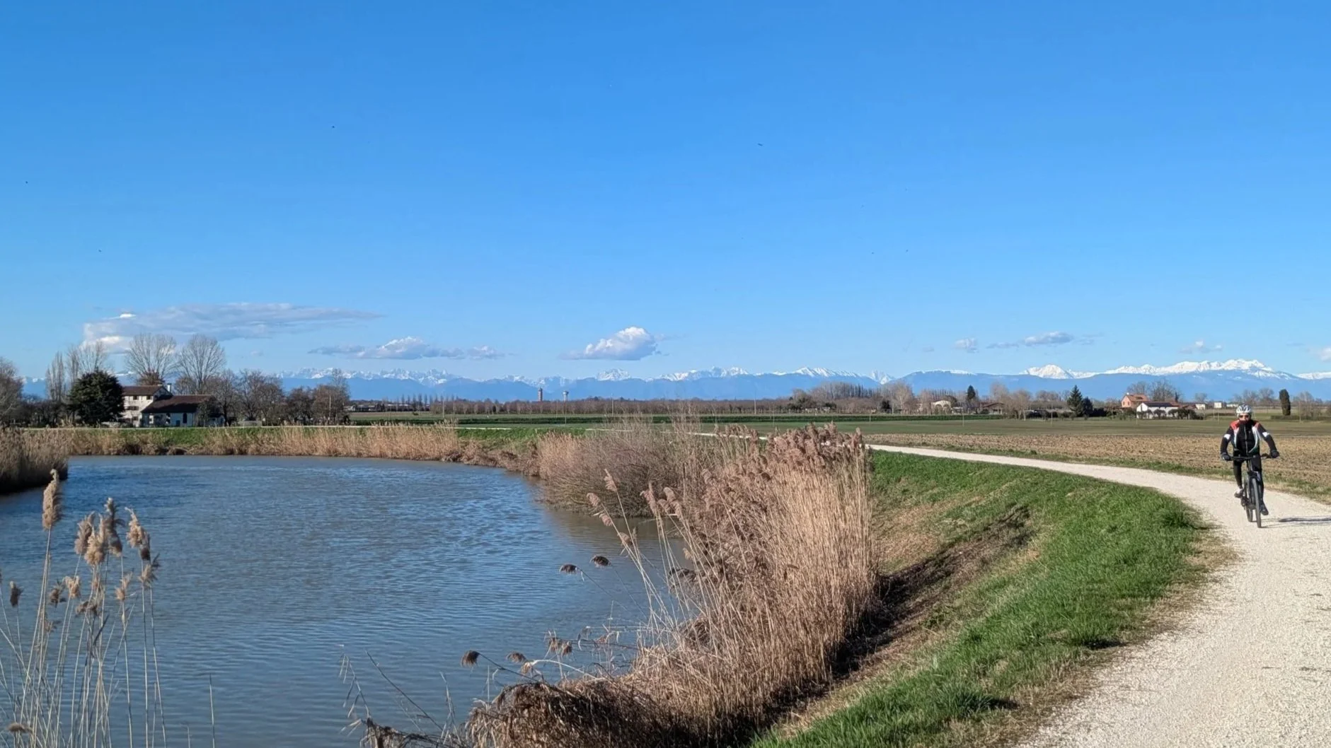 A cyclist riding a path beside a river in a rural area with snow-capped mountains in the background under a clear blue sky.