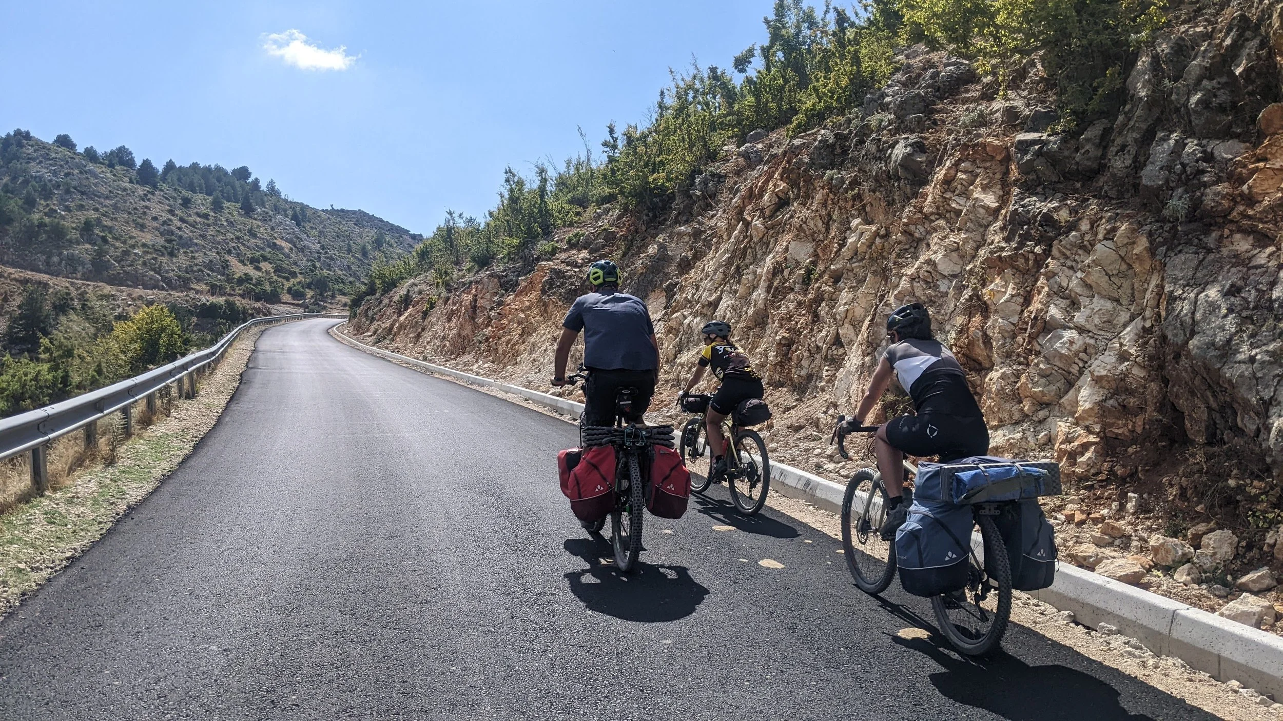 Three cyclists riding on a mountain road with rocky hillside on the right side and a guardrail on the left, under a blue sky with a few clouds.