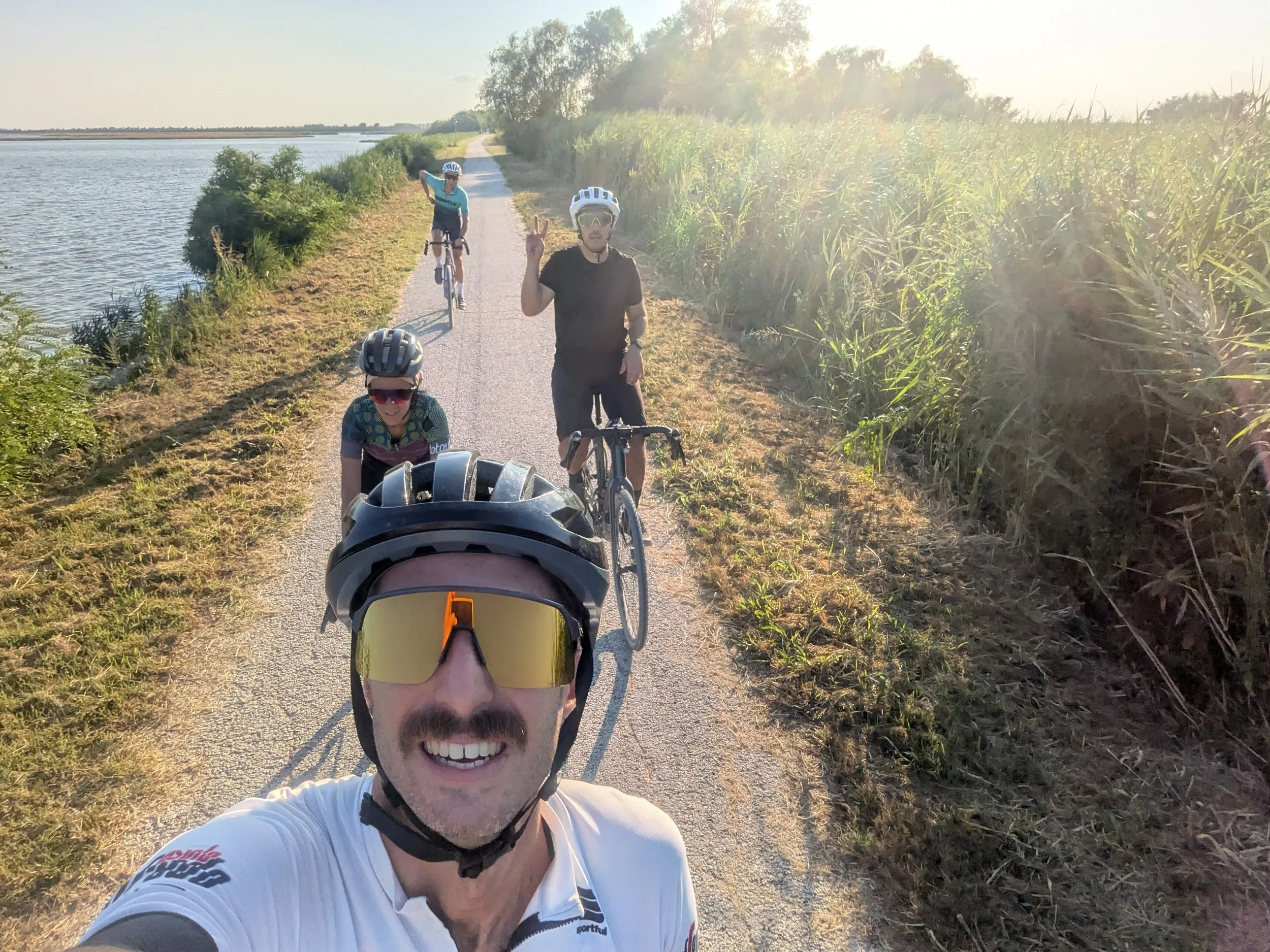 Group of cyclists taking a selfie on a scenic bike trail next to a body of water with tall green grass on the side, sunny weather