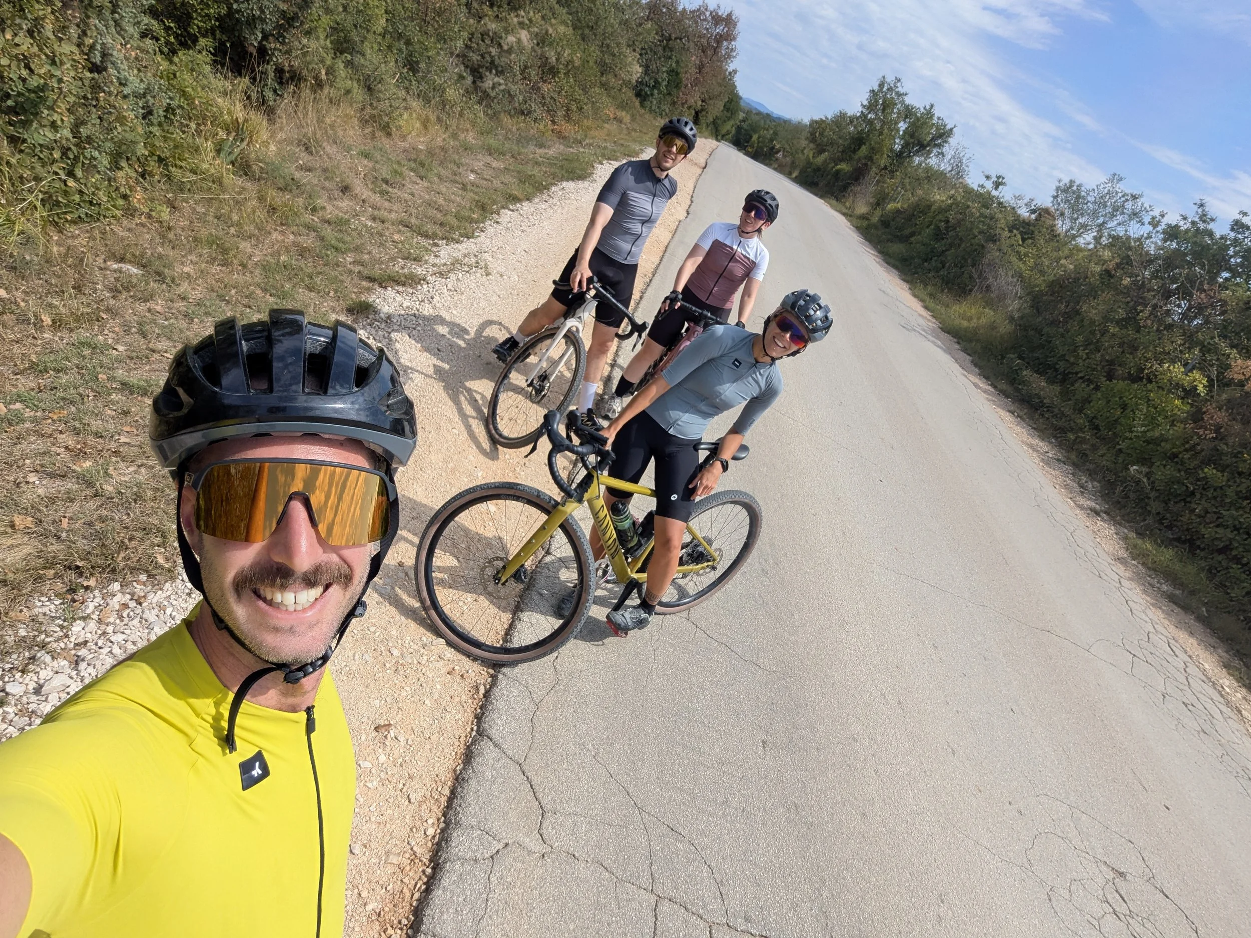 A man in yellow cycling gear with sunglasses taking a selfie with three other cyclists on a rural road surrounded by trees and hills.