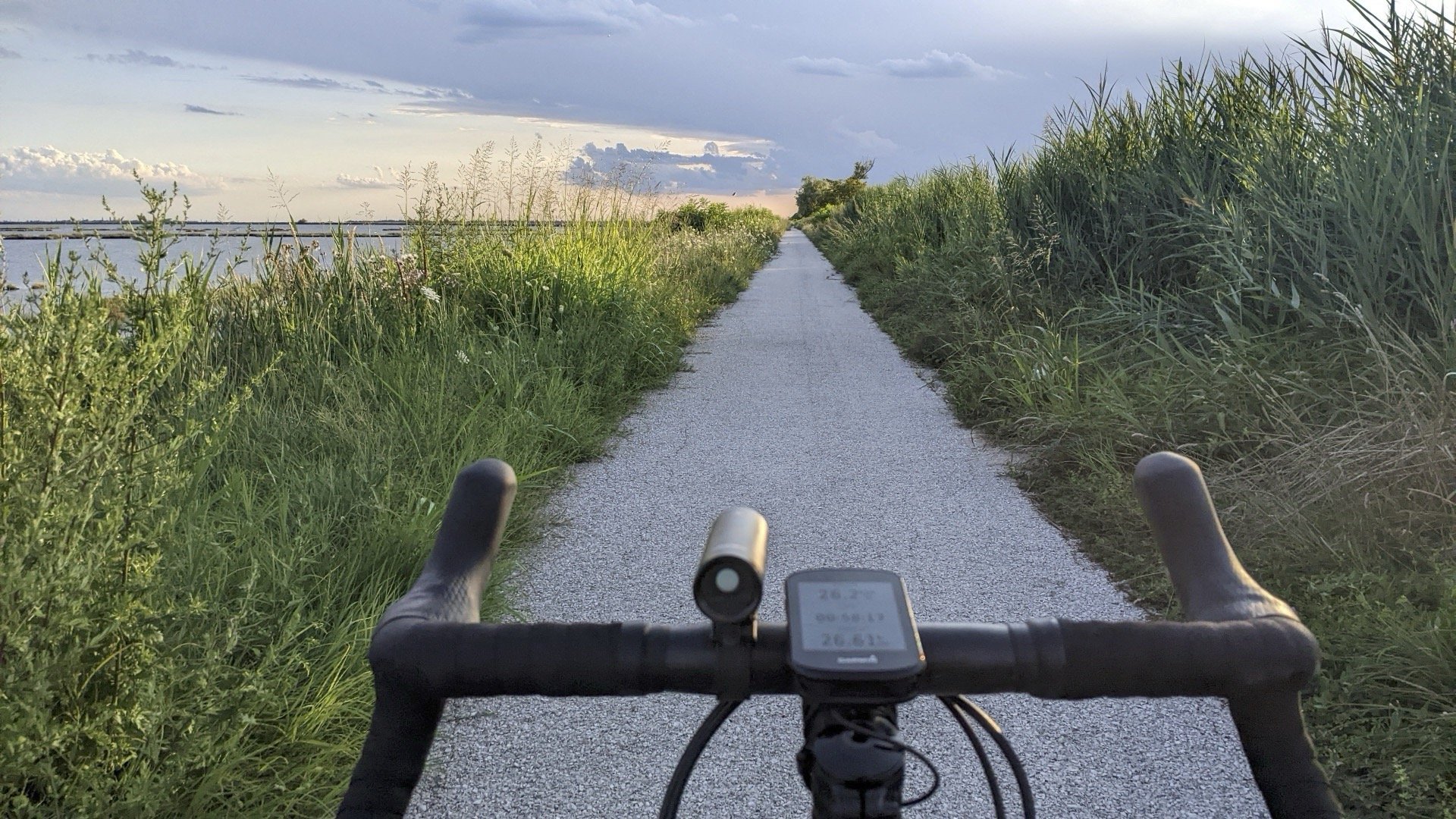 View from a bicycle's handlebars on a narrow gravel trail through tall green grass, with water and a cloudy sky in the distance during sunset.