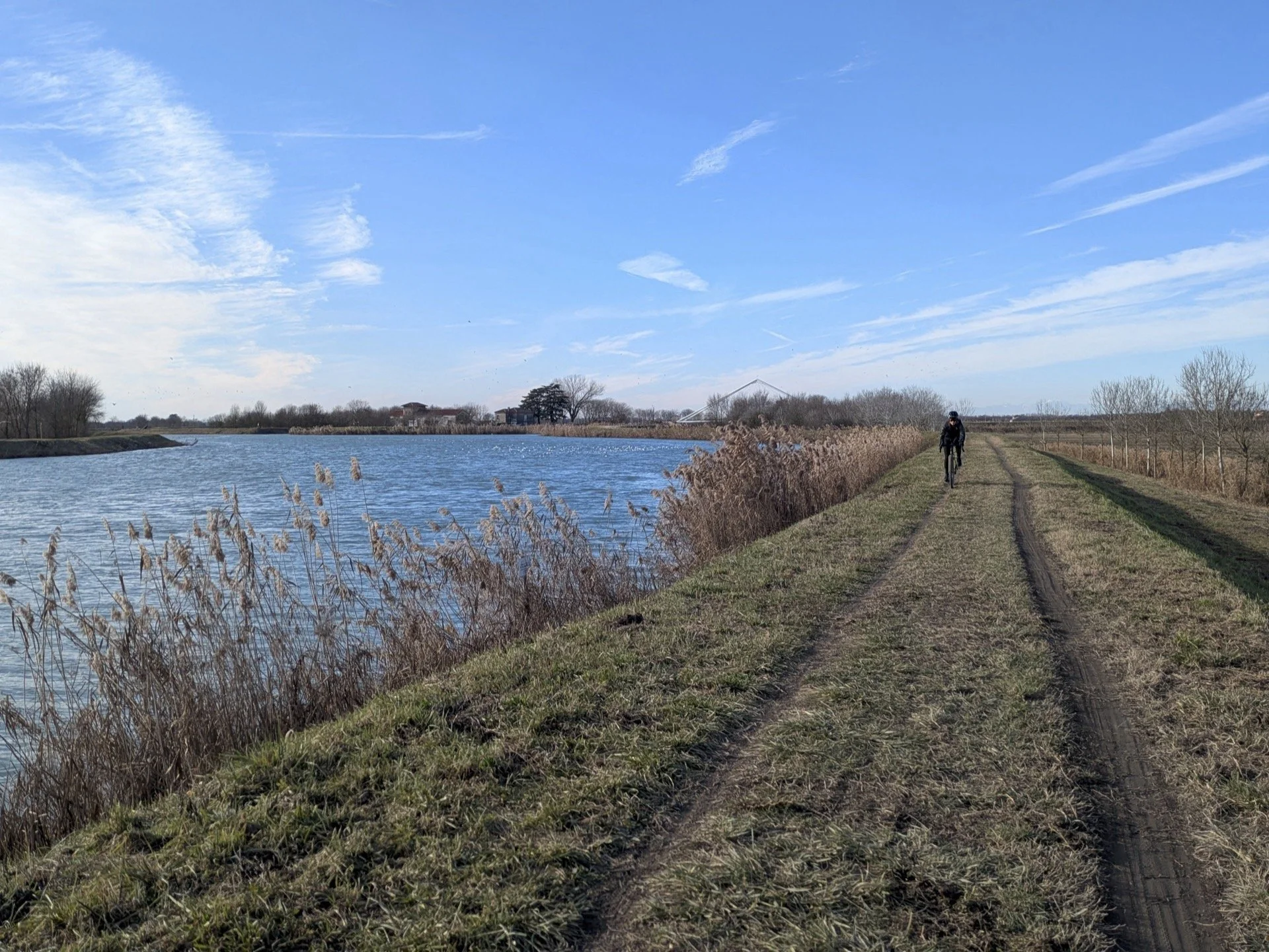 Person riding a bicycle along a grassy trail beside a body of water under a blue sky with scattered clouds.
