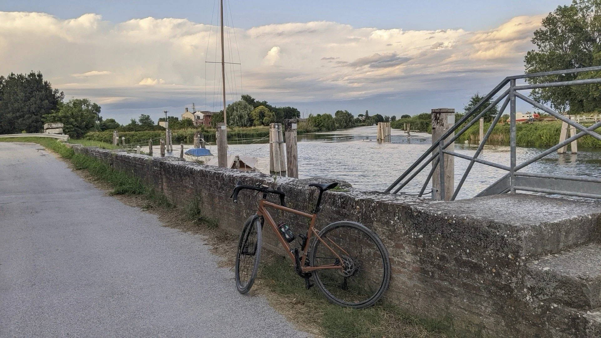 A brown bicycle leaning against a brick wall by a river, with boats and green trees in the background under a partly cloudy sky.