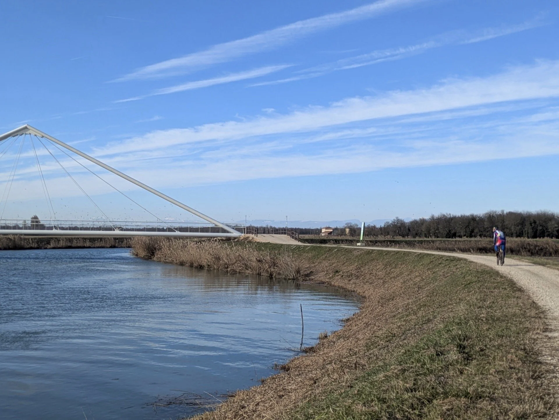 A person riding a bicycle along a dirt path beside a river, with a modern bridge structure in the background, under a clear blue sky with wispy clouds.