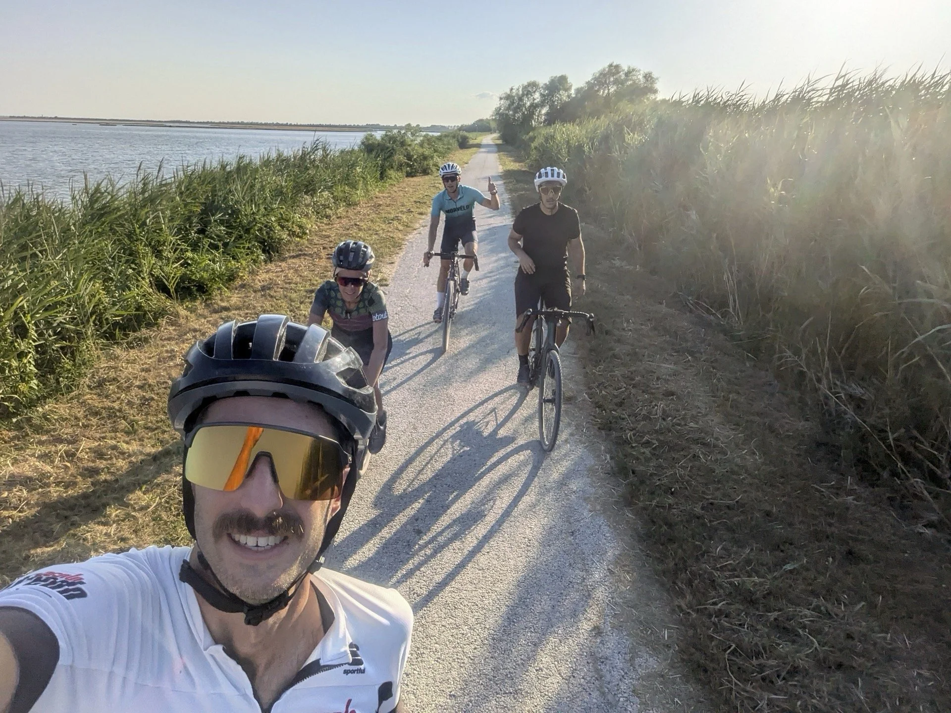 A group of four cyclists taking a selfie on a dirt path beside a body of water, with greenery on both sides and the sun shining brightly.