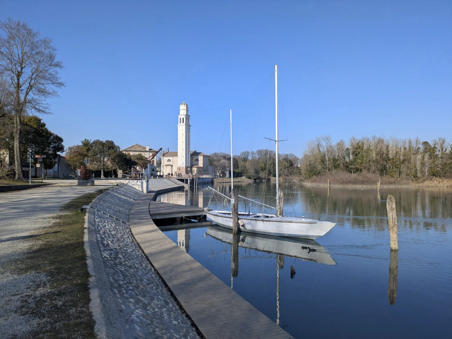 A peaceful river scene with a white sailboat docked near a curved walkway, a tall clock tower in the background, and trees along the shoreline under a clear blue sky.