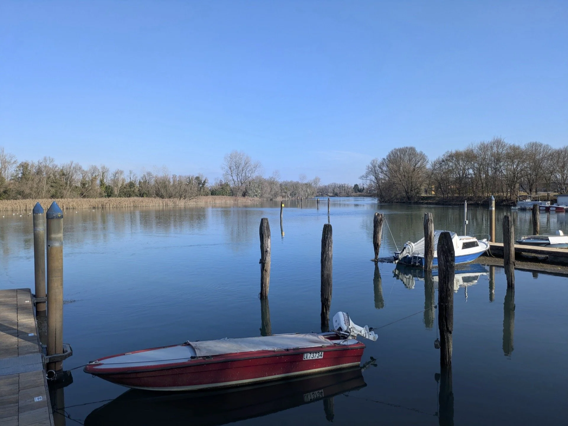 A calm river with boats docked at a pier, surrounded by trees under a clear blue sky.