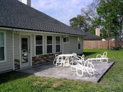 Backyard patio with a table and chairs in front of a house with a grey roof and siding, fenced yard, and green grass.