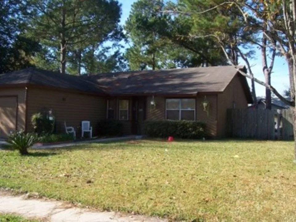 A single-story brown house with a lawn in front, two white chairs near the entrance, trees in the background, and a wooden fence on the right side.