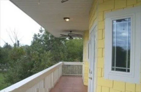 A view of a balcony with a yellow house exterior, white window, railing, and trees in the background.