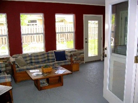 Living room with red walls, three windows, a glass door, a plaid sofa with pillows, a wooden coffee table with magazines and a basket, and a view of a fenced backyard.