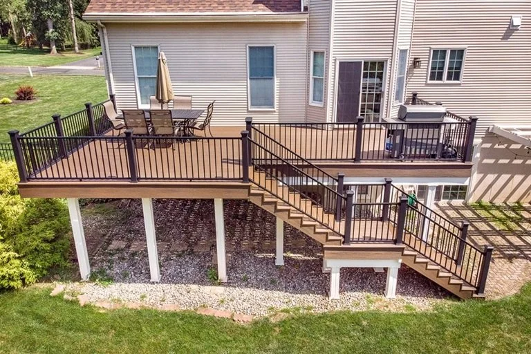 Backyard with wooden deck and metal railing stairs leading down to grass and gravel area, attached to a beige house with multiple windows and sliding glass door.