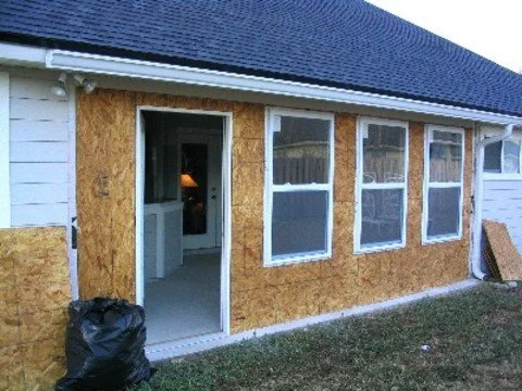 Side of a house under construction with OSB sheathing, multiple windows, and an open door, with a black trash bag on the ground.